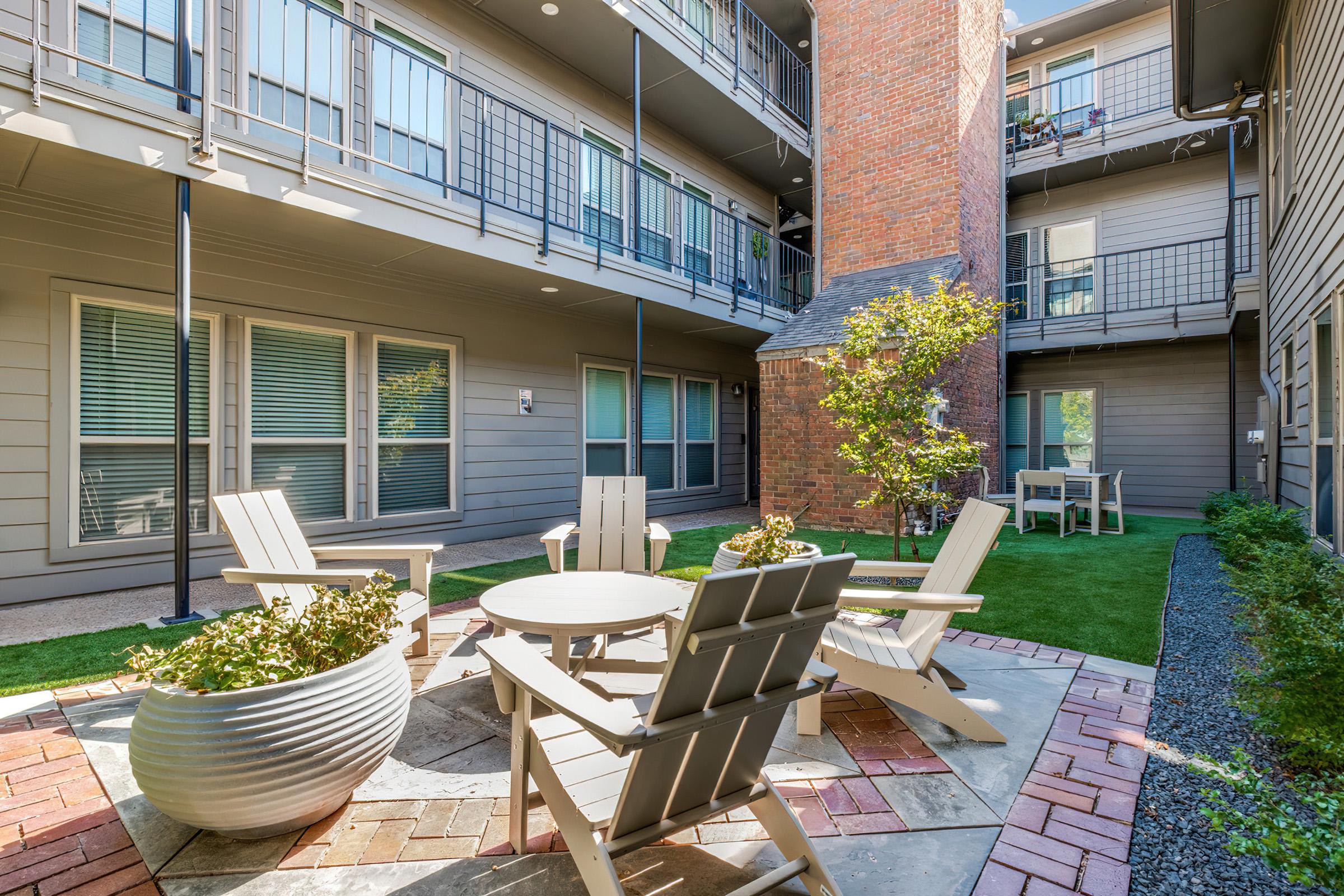 A communal outdoor area featuring a circular table surrounded by several wooden chairs. There is a small tree and greenery in the corner, with brick pathways and a modern apartment building in the background, creating a cozy and inviting atmosphere.