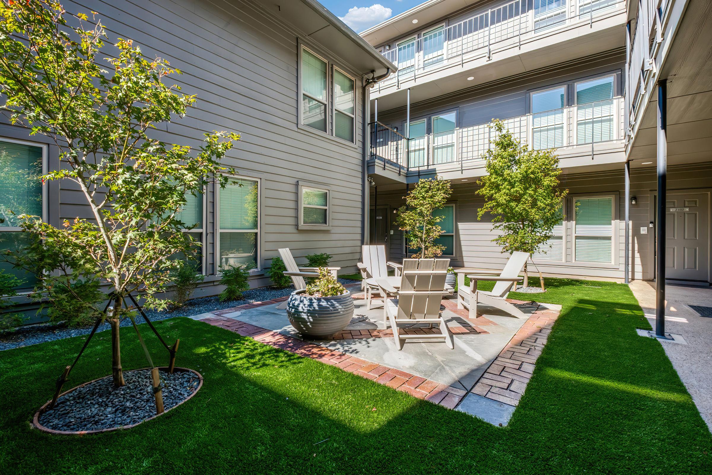 A well-maintained courtyard featuring a seating area with light-colored chairs and a table, surrounded by greenery. There are potted plants and trees, with a mix of grass and stone pathways, situated between residential buildings. Natural light illuminates the space, creating a welcoming atmosphere.