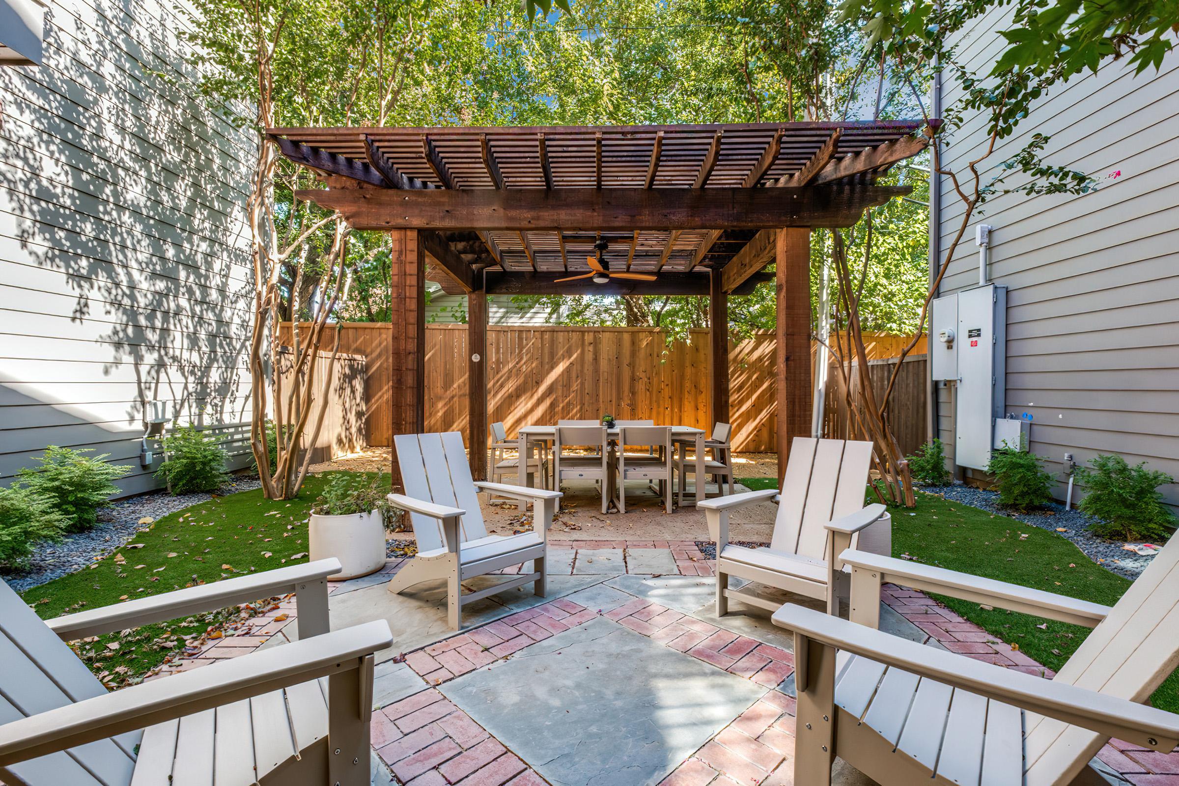 A cozy outdoor space featuring a wooden pergola with a ceiling fan, surrounded by four white lounge chairs and a circular stone and brick patio. Lush greenery and small bushes create a serene atmosphere, ideal for relaxation or gatherings.