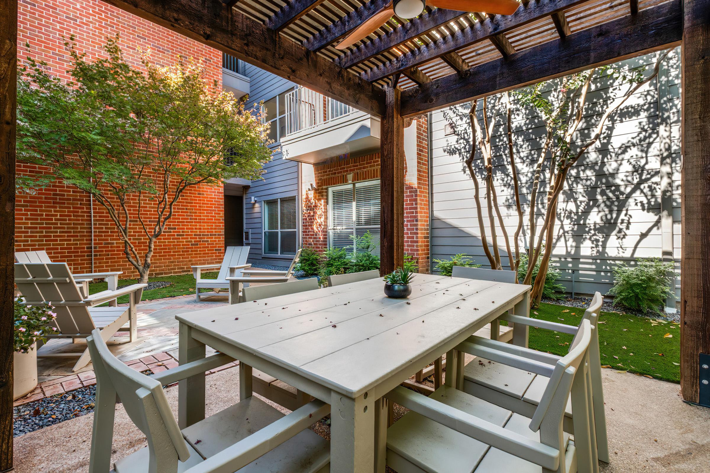 A peaceful outdoor patio area with a wooden pergola, featuring a large white table surrounded by matching chairs. Lush greenery, including trees and shrubs, frames the space, creating a cozy atmosphere. The backdrop includes brick and siding of nearby buildings, enhancing the charming setting.