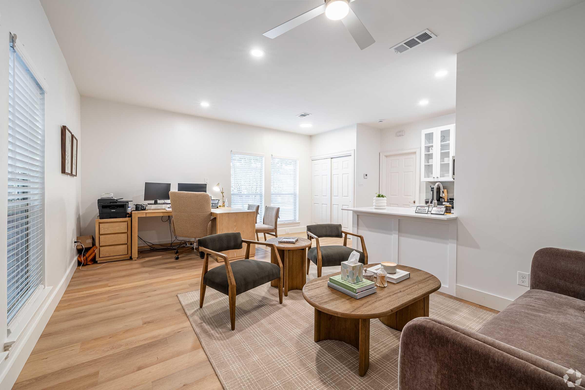 A modern living space featuring a cozy seating area with brown chairs and a sofa, a wooden coffee table, and a desk with a computer in the background. The room has large windows allowing natural light, light-colored walls, and wooden flooring, creating a warm and inviting atmosphere.