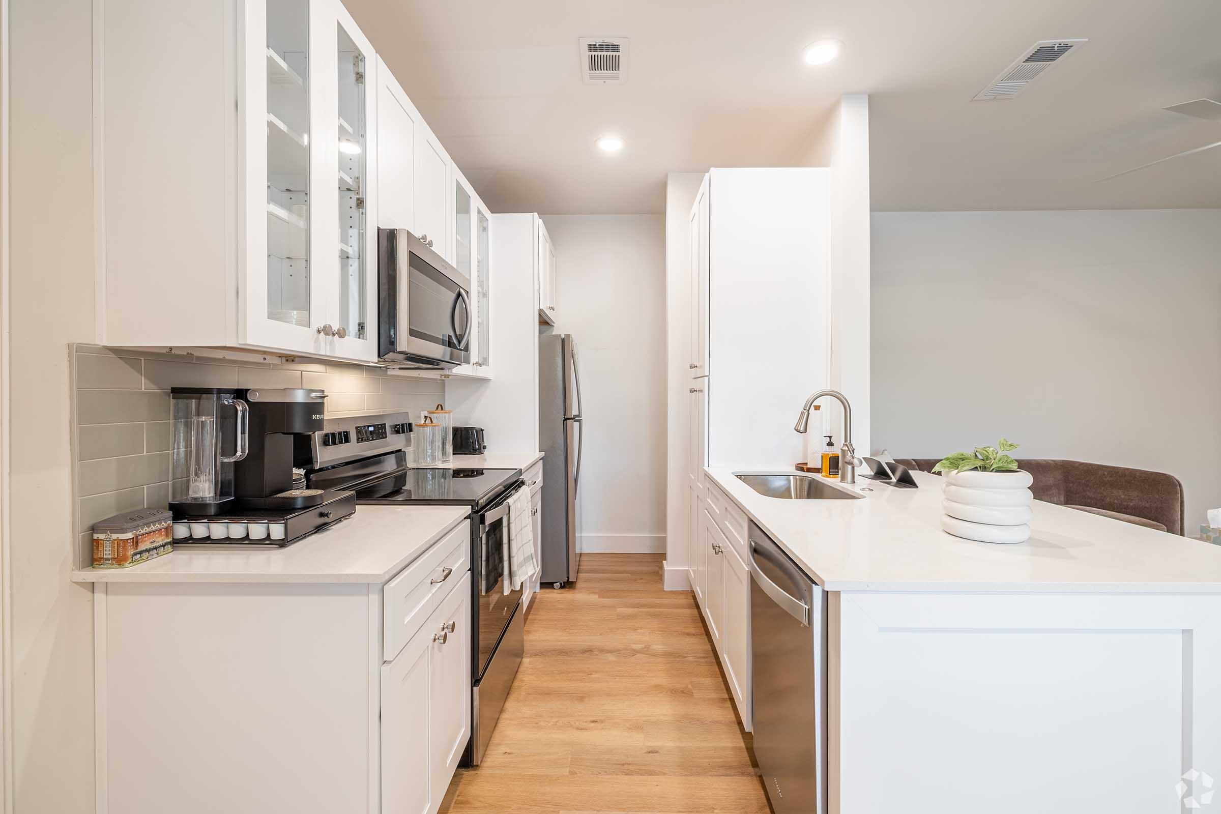 A modern kitchen featuring white cabinets, stainless steel appliances including a microwave and refrigerator, and a sleek island with a sink. The countertops are light-colored, and the flooring is wooden. Soft lighting creates a bright and inviting atmosphere.