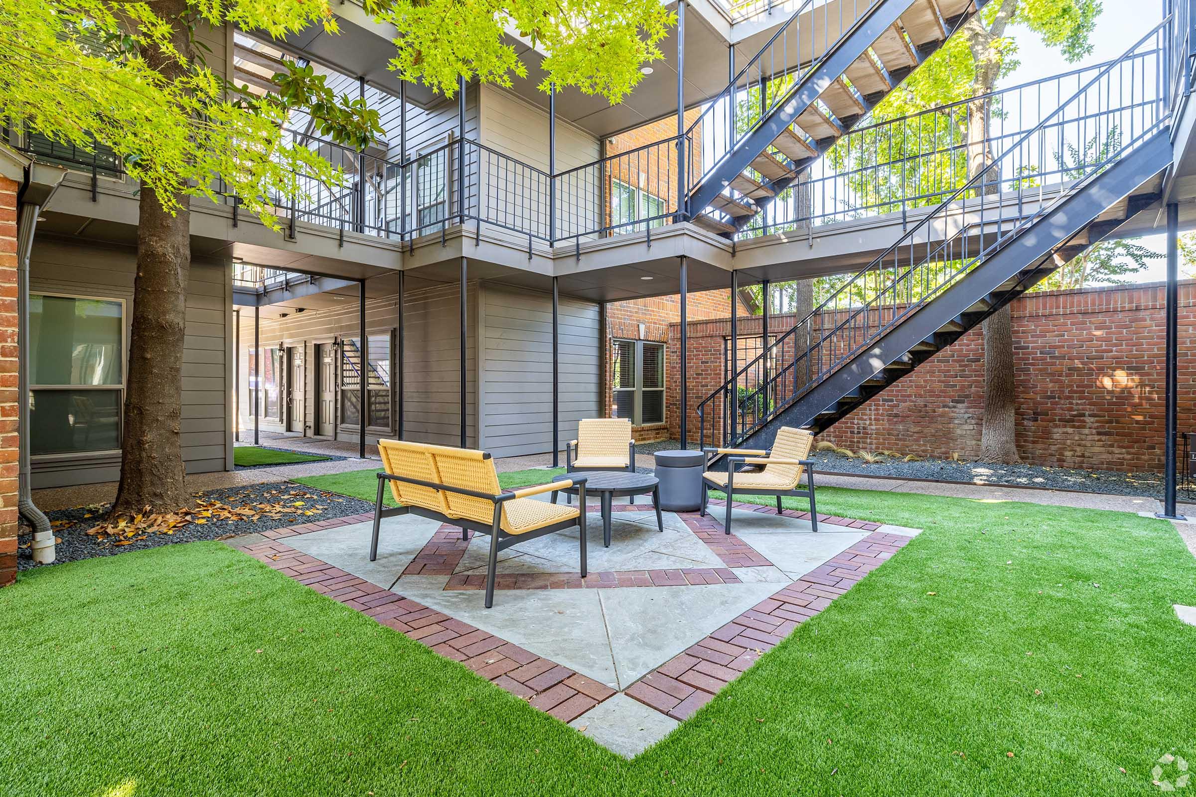 A communal outdoor courtyard featuring modern seating arrangements with benches around a central fire pit, surrounded by greenery and brick walls. A staircase leads to upper floors, and lush grass covers the ground, creating a welcoming atmosphere for relaxation and social gatherings.