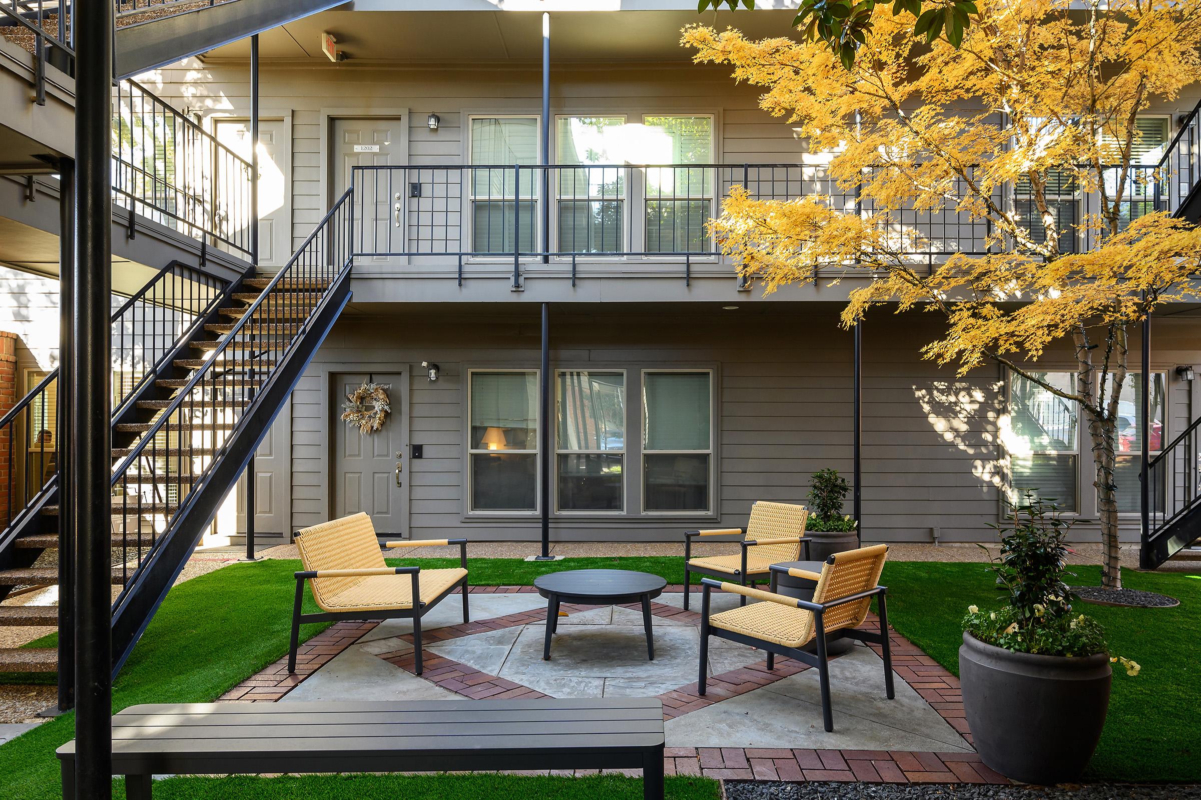 A cozy outdoor courtyard featuring a round fire pit surrounded by four yellow chairs, with green grass and a decorative tree. In the background, there are two levels of apartments with balconies and a welcoming wreath on one door, creating an inviting and relaxing atmosphere.
