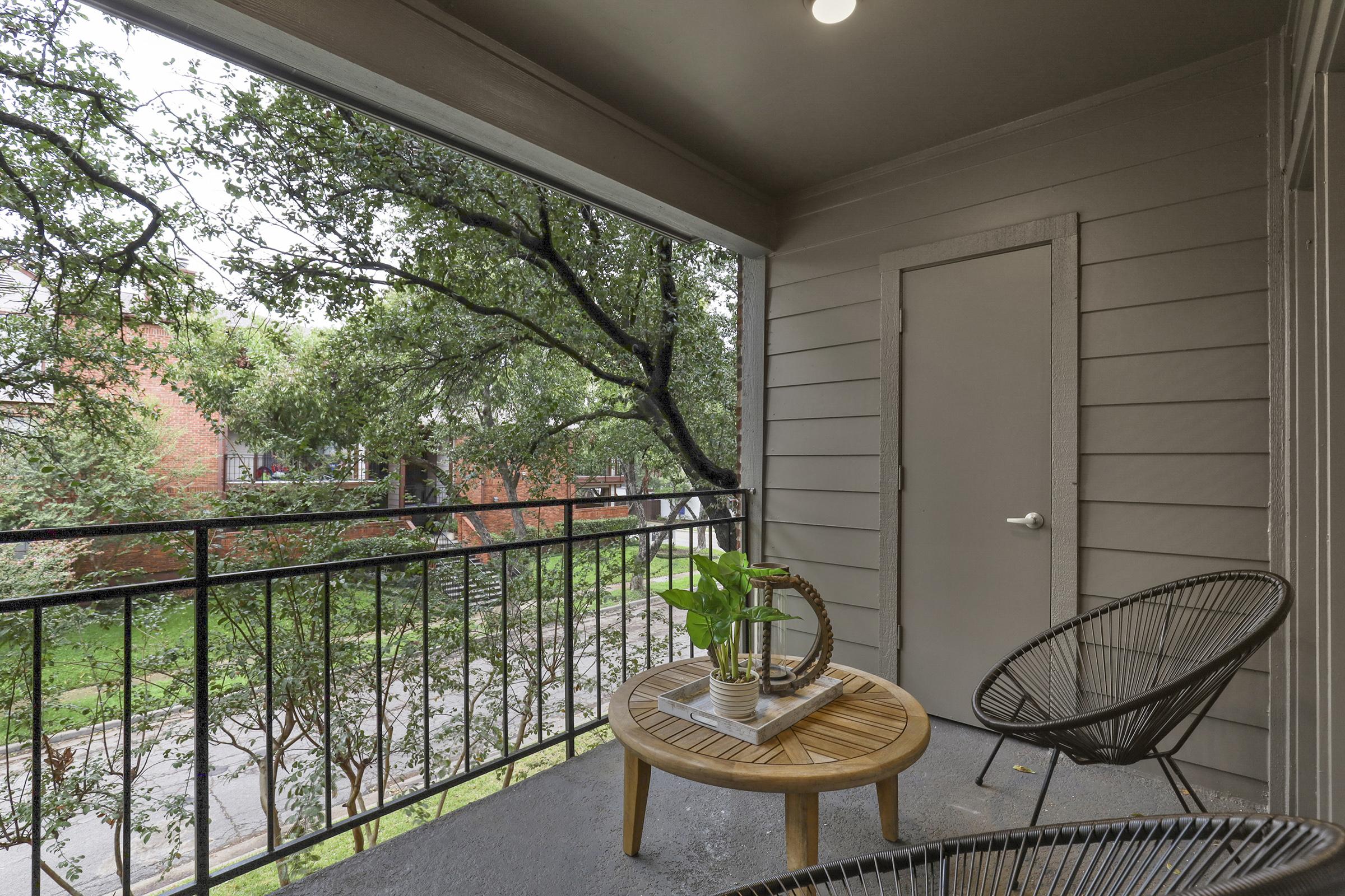 A cozy balcony featuring a circular wooden table with a potted plant and decorative items. Two stylish black chairs surround the table, and greenery is visible in the background, along with a closed door and a railing framing the view of the street below.