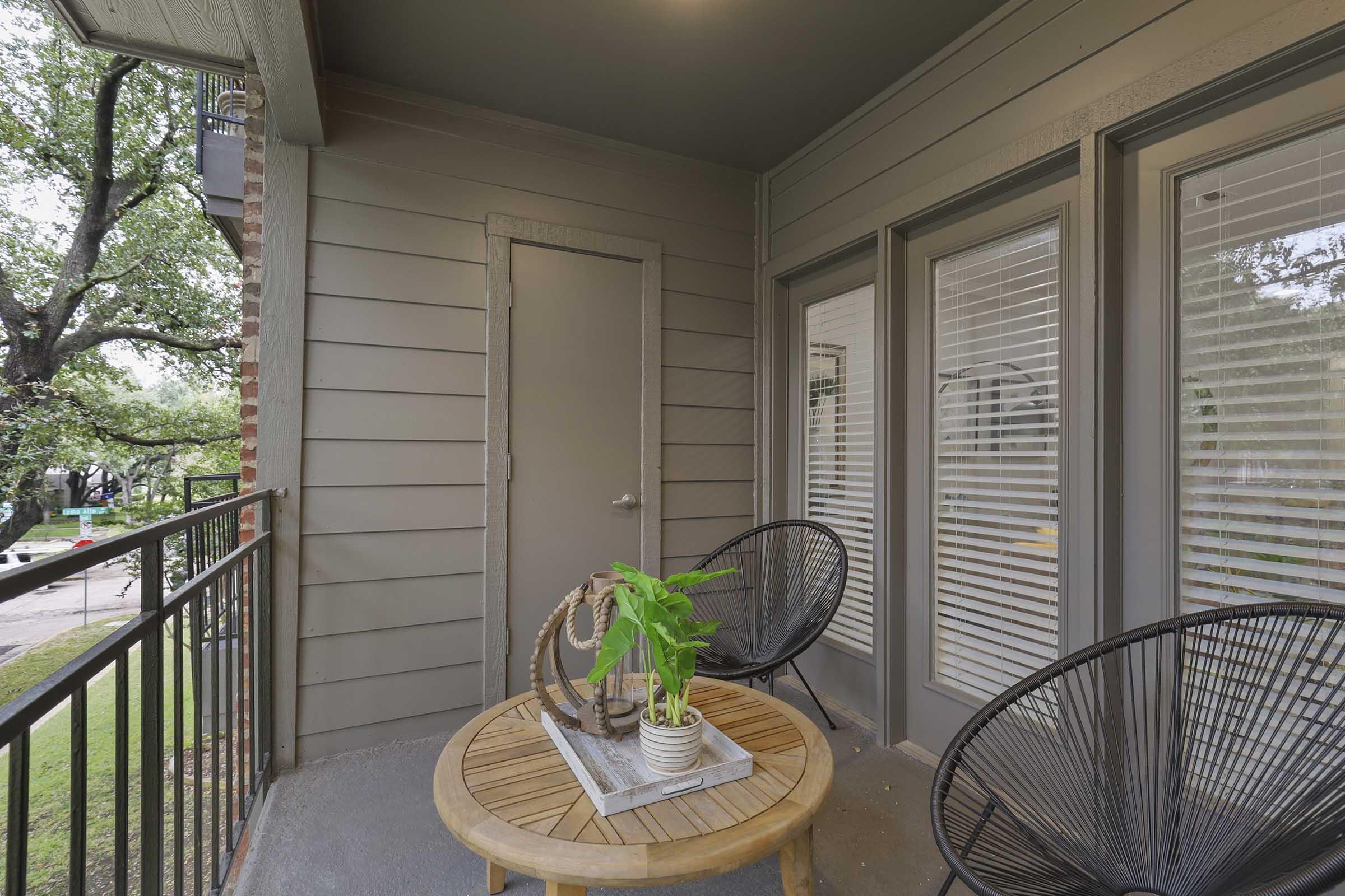 A cozy balcony featuring two black wicker chairs and a round wooden table. A small potted plant sits on the table, and there is a closed door visible. Sunlight filters through the nearby trees, creating a serene outdoor space. The walls are painted in a soft gray tone, enhancing the inviting atmosphere.