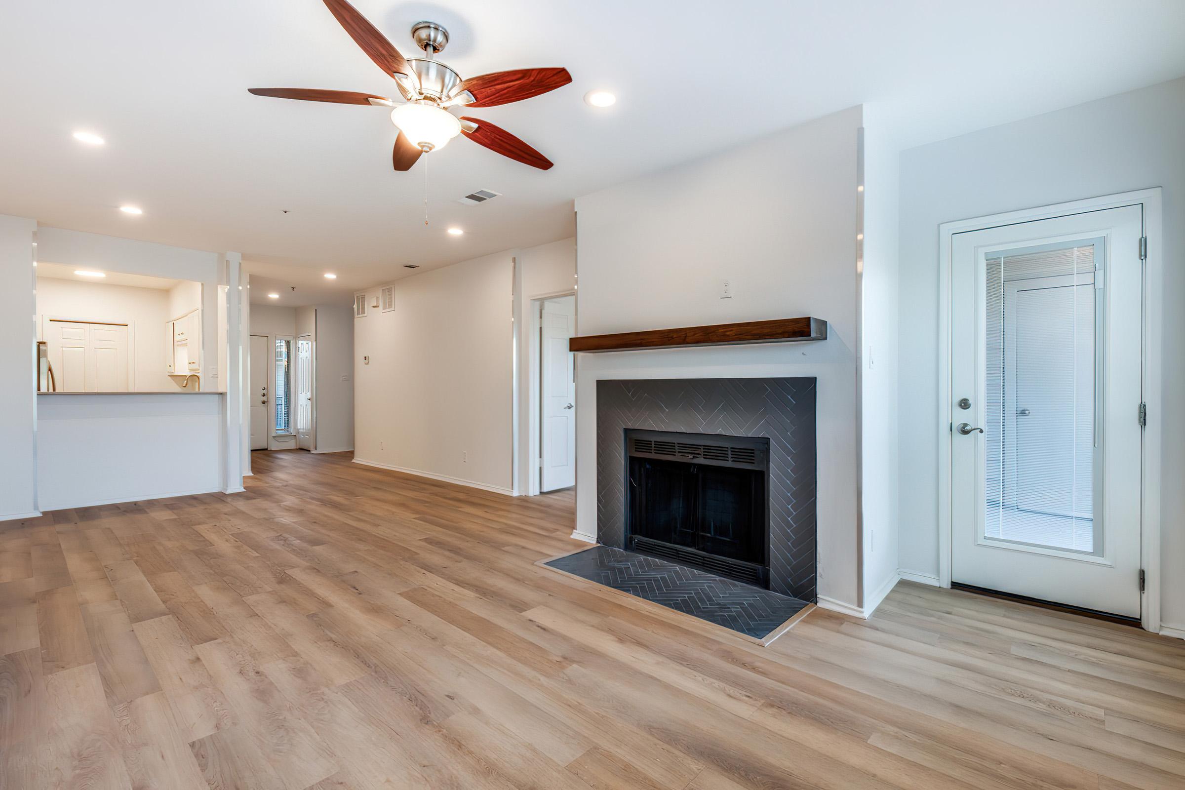 A spacious living room featuring light hardwood flooring, a ceiling fan, and a modern fireplace with a herringbone pattern. The room is well-lit with natural light, and there is an entrance to a patio on the right. A hallway leads to the kitchen area. The walls are painted in soft neutral tones.