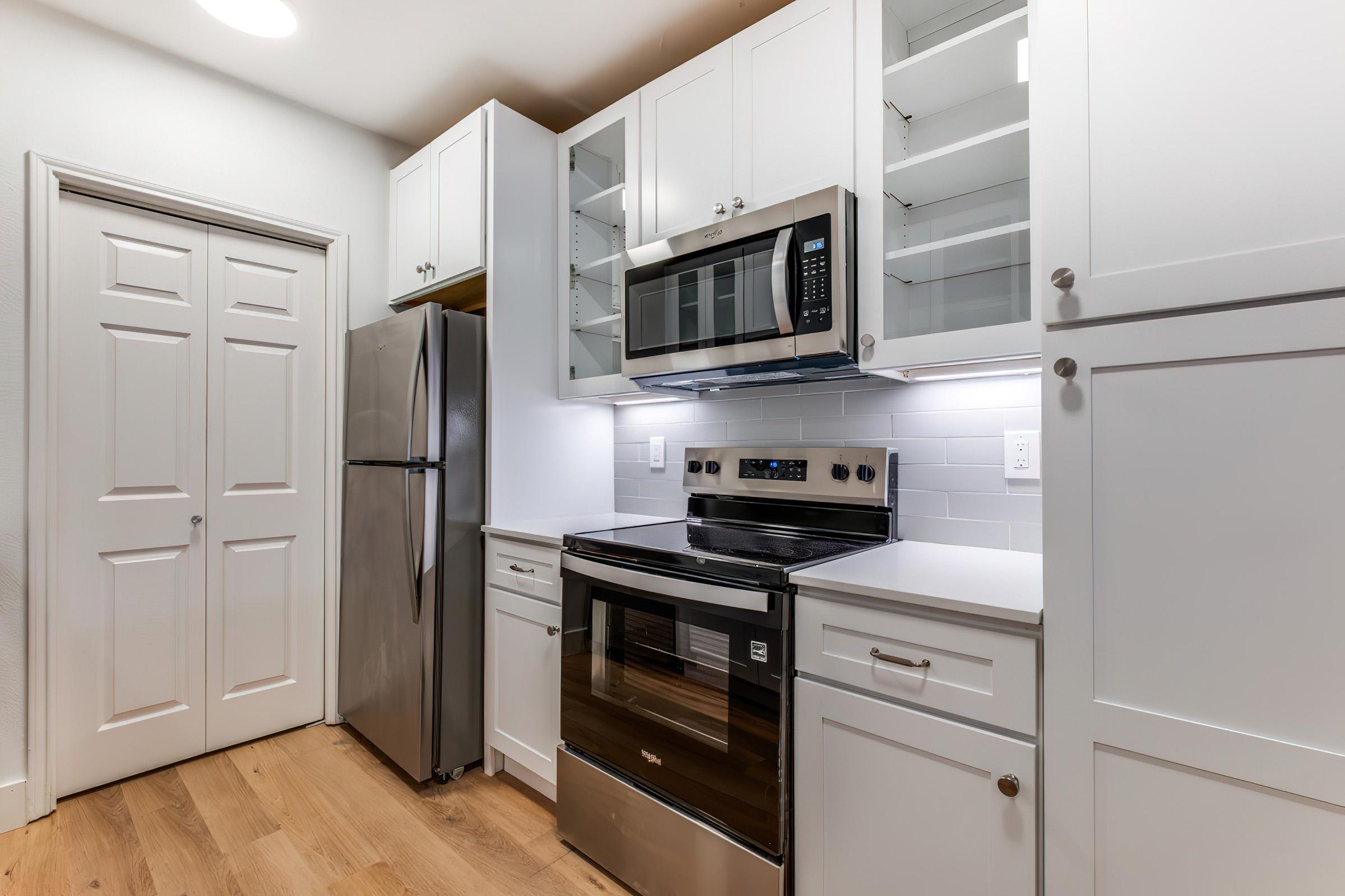 Modern kitchen featuring white cabinetry, stainless steel appliances, including a microwave and an oven, alongside a refrigerator. The countertops are light-colored, and the space has wooden flooring. Glass-front cabinets are empty, allowing for a bright and open feel.