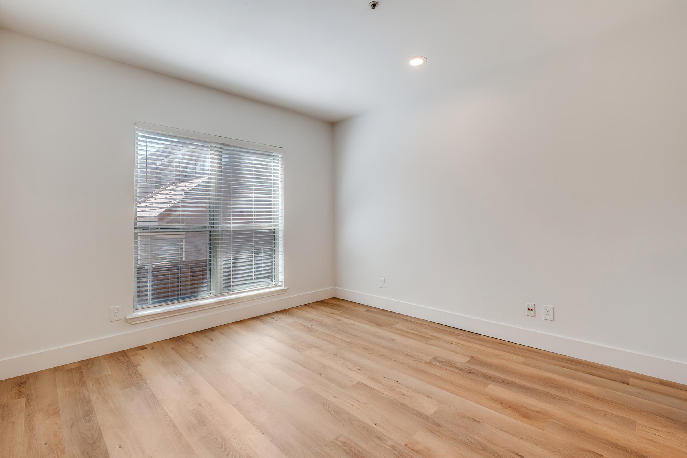 A bright, empty room with light wood flooring and neutral-colored walls. A large window with white blinds allows natural light to fill the space. The room is uncluttered, providing a blank canvas for potential furniture arrangement or decoration.