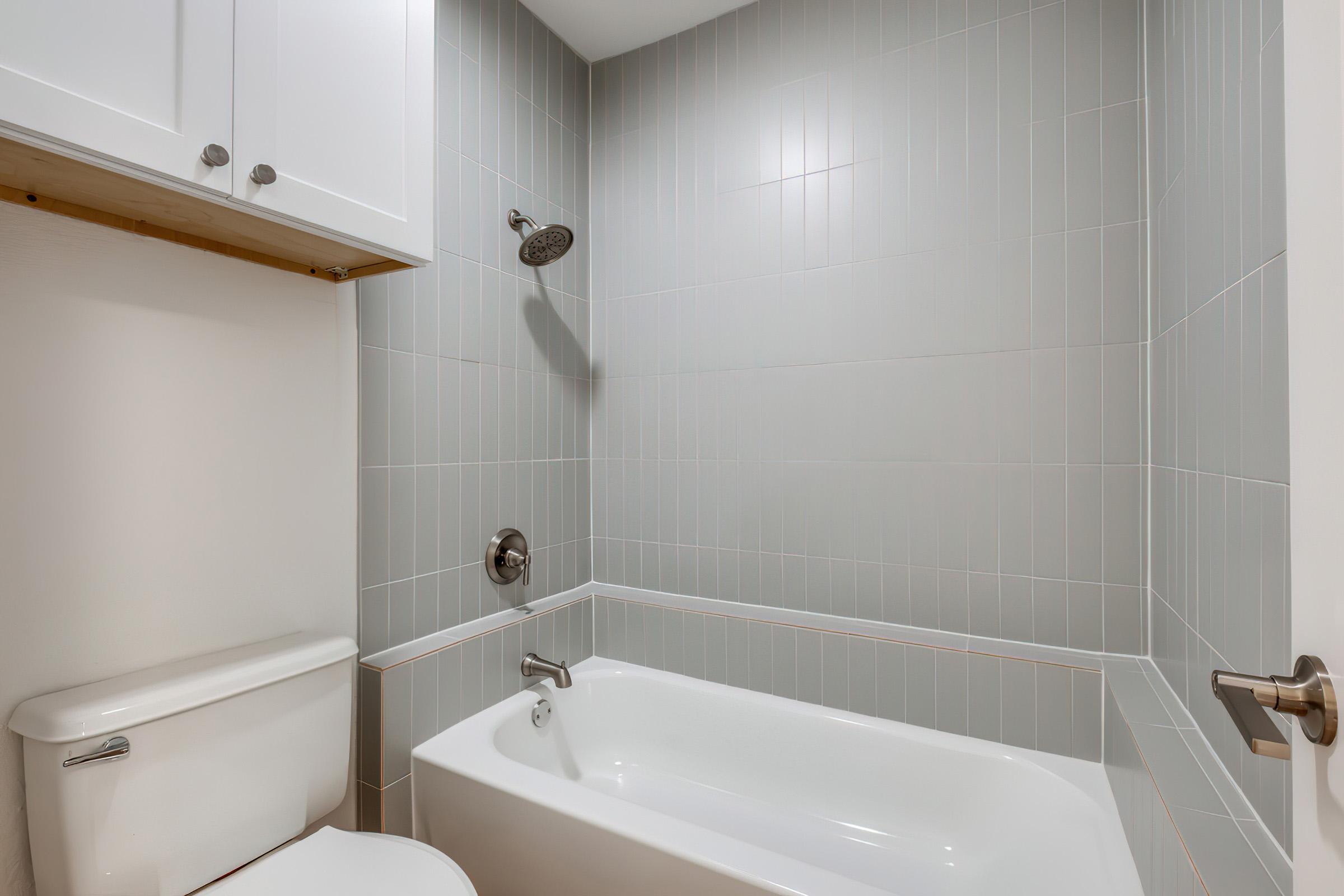 A modern bathroom featuring a white bathtub against gray tiled walls. A chrome showerhead is mounted above the tub. To the left, there is a white toilet, and above the tub, a wooden shelf is installed under white cabinets. The overall design is minimalist and clean.