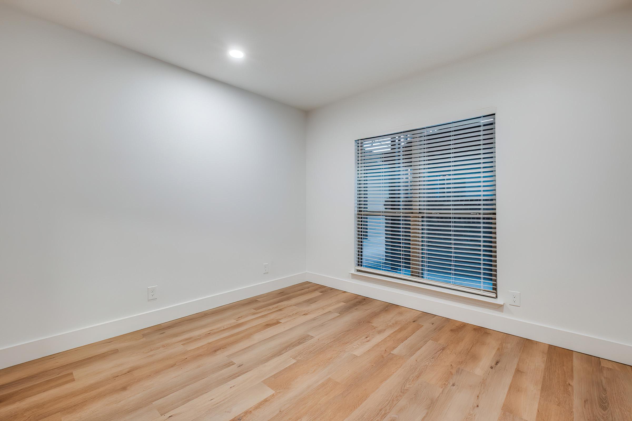 Empty room with light-colored walls and wooden flooring. A window with blinds allows natural light to enter. The space is minimalistic and uncluttered, creating a serene atmosphere.