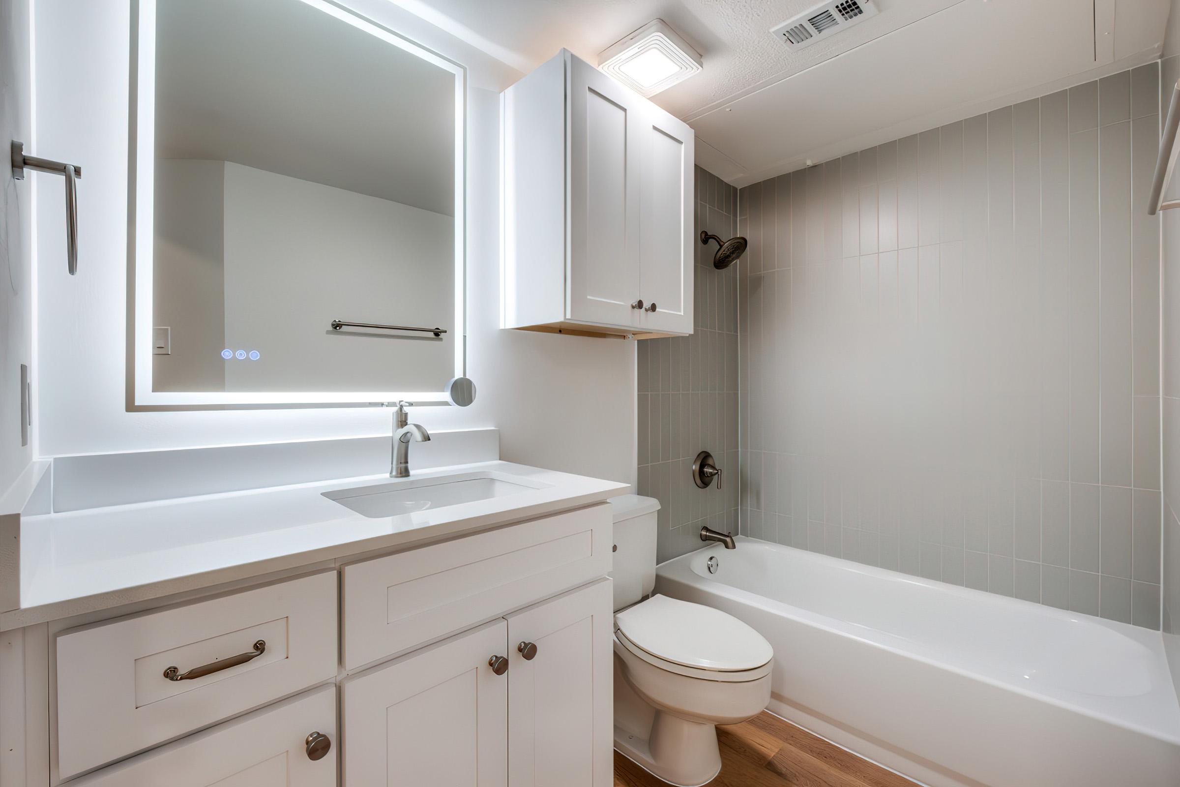 A modern bathroom featuring a white vanity with a rectangular mirror and illuminated edges, a single faucet, a bathtub with a showerhead, a toilet, and grey tiled walls. The floor is wood-like. Natural light illuminates the space, creating a clean and inviting atmosphere.
