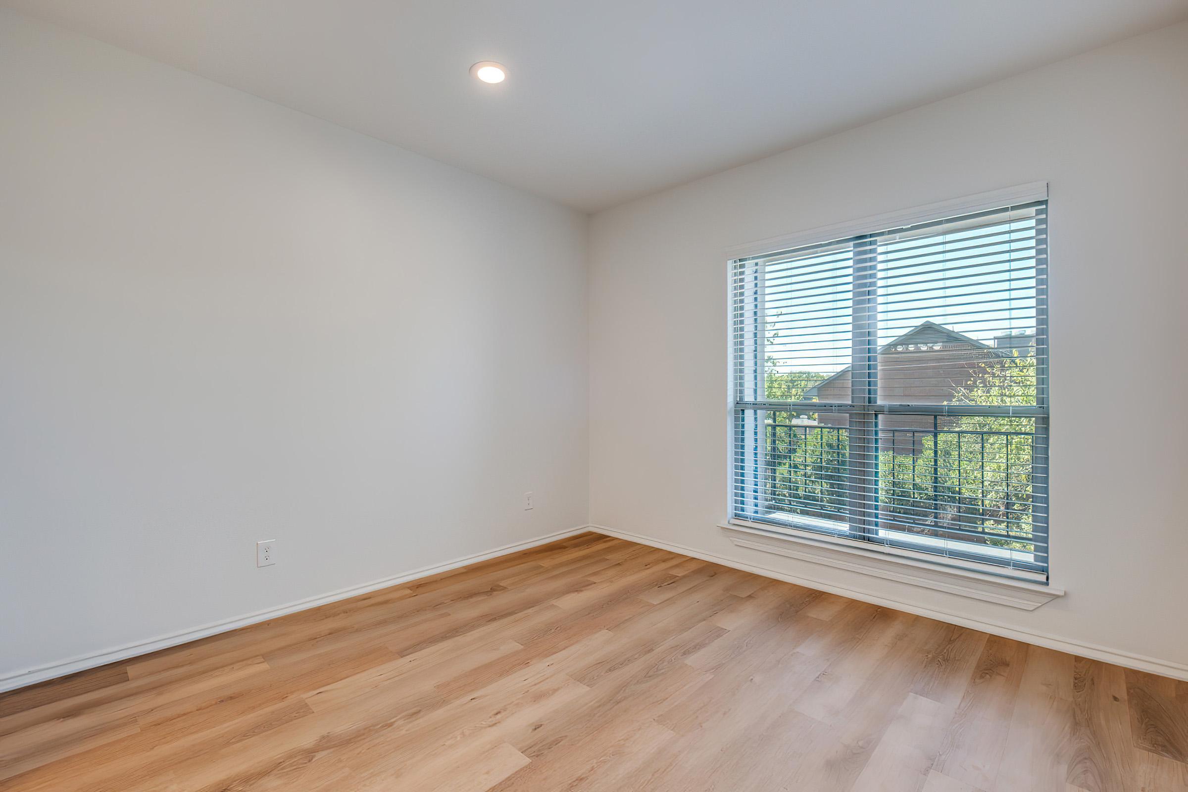 A bright, empty room with white walls and wooden flooring. There's a large window with blinds allowing natural light to illuminate the space, and a view of greenery outside. The room has a clean and modern feel, perfect for various uses.