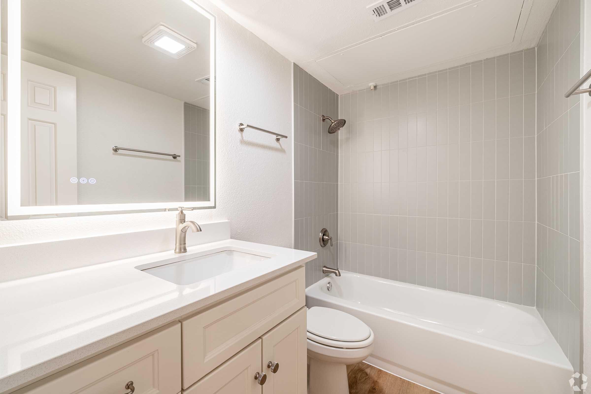 A modern bathroom featuring a white sink with a silver faucet, a mirror above the sink, light gray tiled walls, a bathtub with a shower, and a compact toilet. The space is well-lit and has a clean, minimalist design.