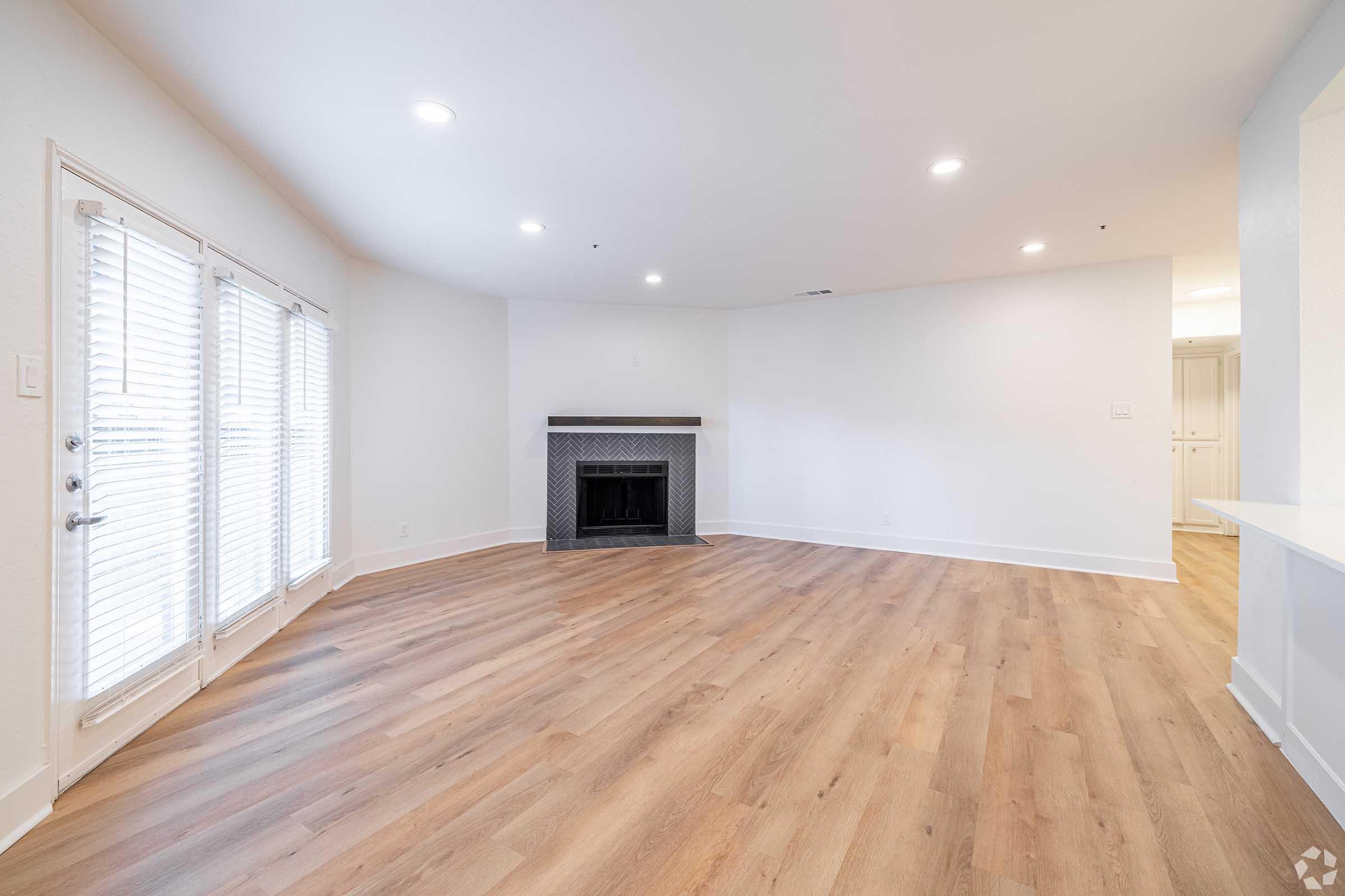 A spacious, empty living room featuring light wood flooring, a fireplace with a tiled surround, and large windows with white shutters. The walls are painted in a soft, neutral color, creating a bright and inviting atmosphere. A doorway leads to another room in the background.