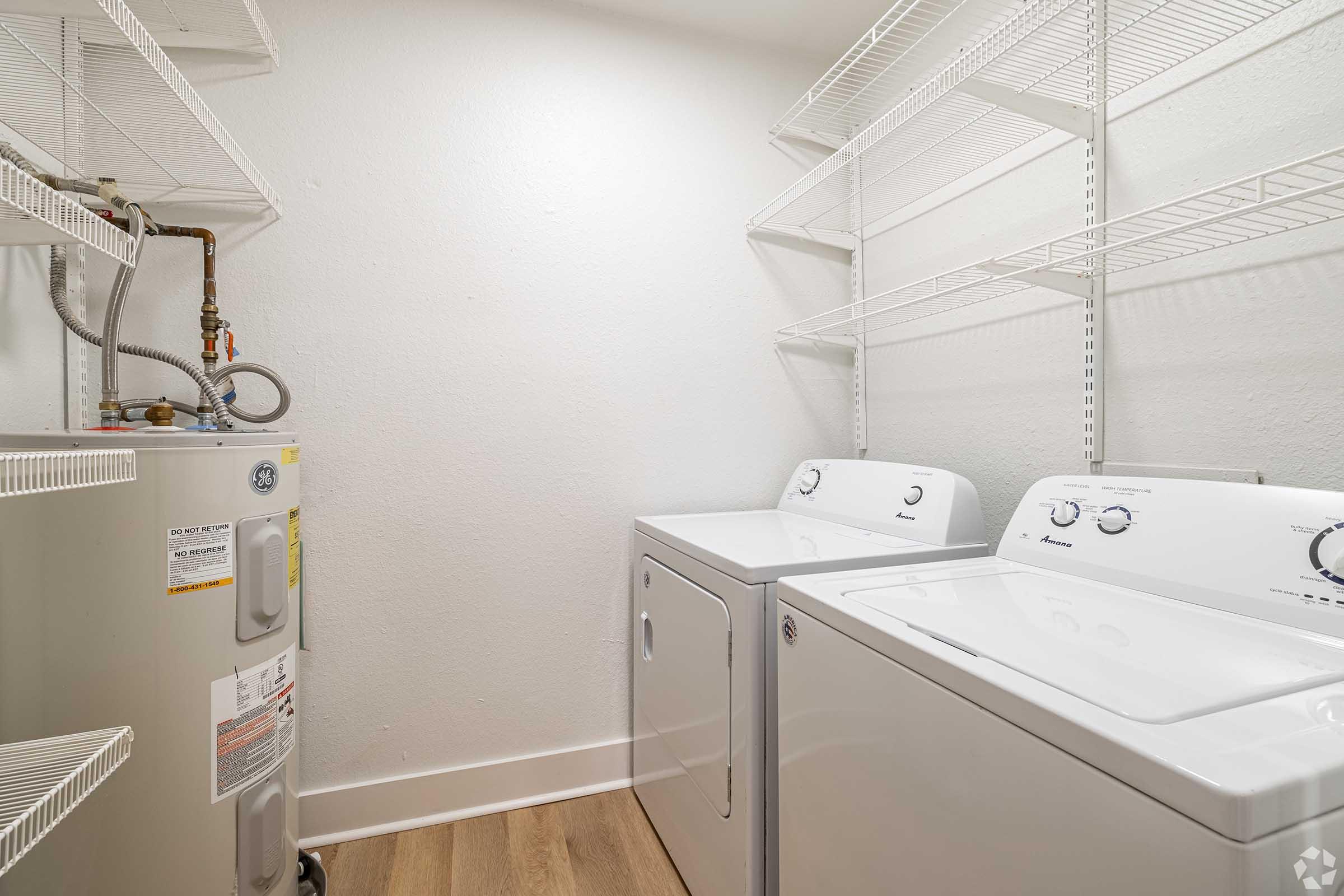 A clean laundry room featuring a white washing machine and dryer side by side. Shelving above the appliances provides storage space. A water heater is located next to the washer, against a lightly colored wall, with wooden flooring adding warmth to the room.