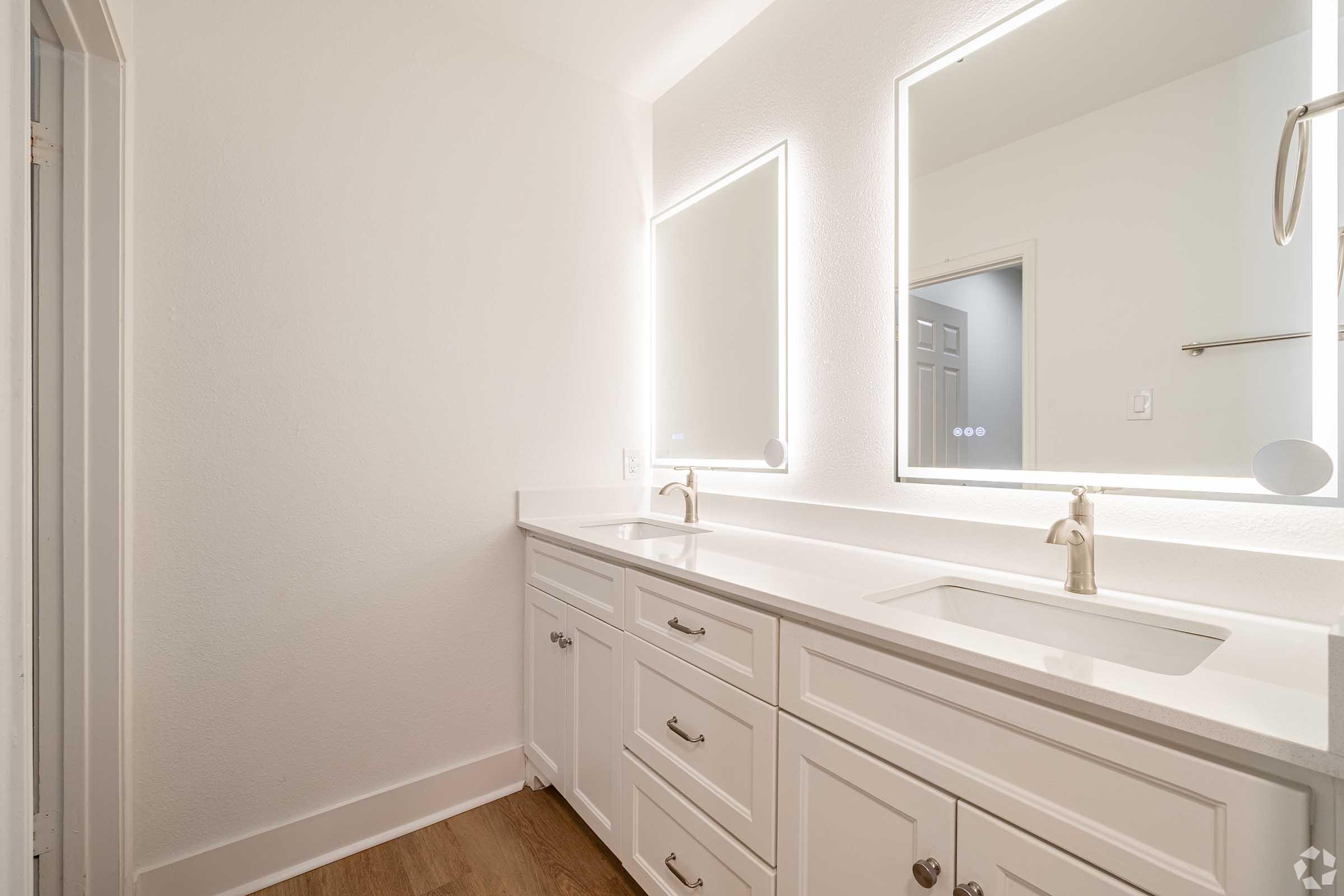 A modern bathroom featuring dual sinks, white cabinetry, and illuminated mirrors. The space has a clean, minimalist design with light-colored walls and wood-like flooring, creating a bright and airy atmosphere. A door leads to another room in the background.