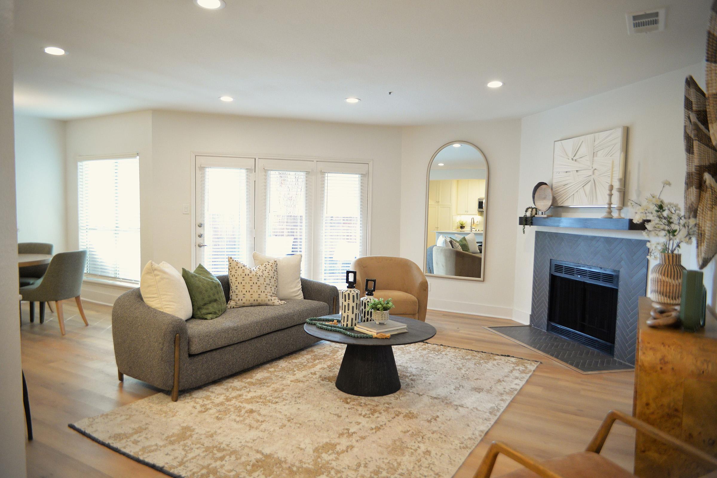 A cozy living room featuring a comfortable gray sofa with decorative pillows, a round black coffee table, and a light-colored area rug. There is a fireplace with a minimalist mirror above it, and large windows letting in natural light. The space also includes a dining area with a small round table and chairs.