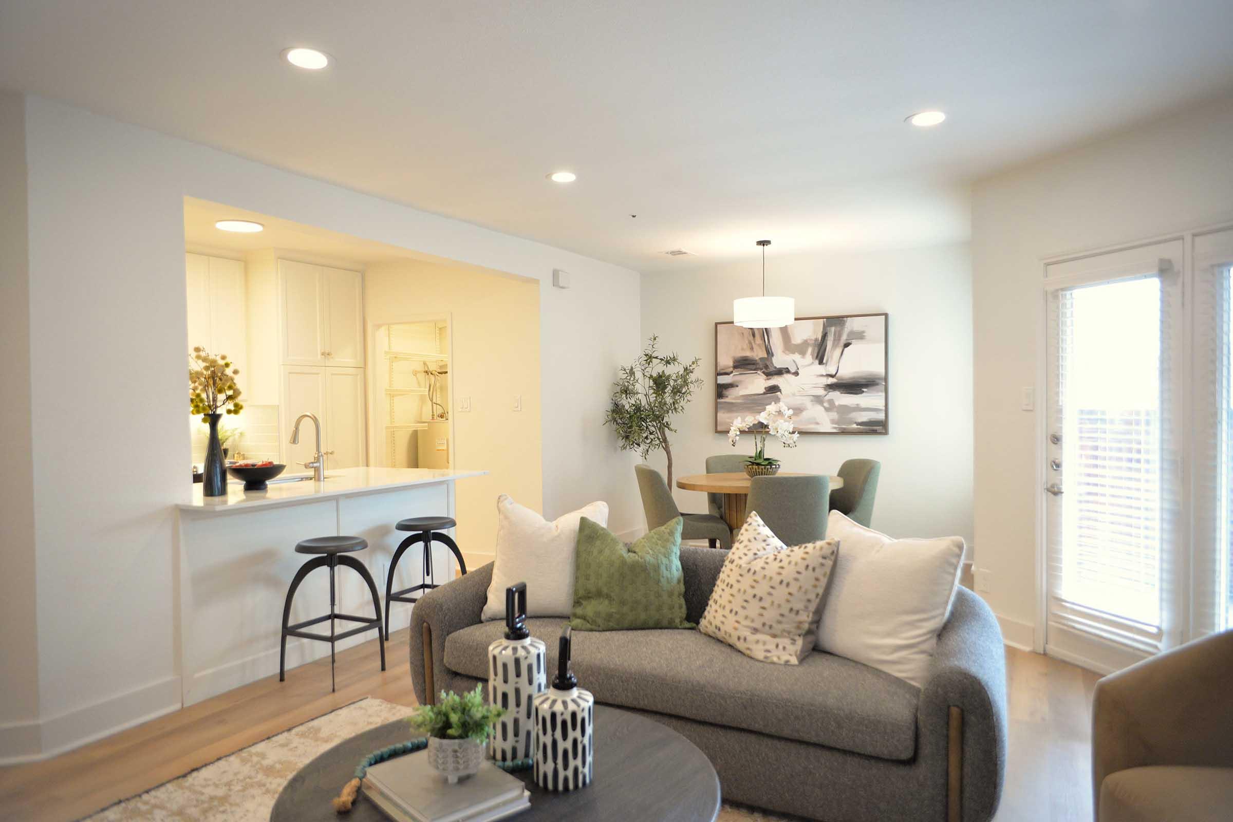 A modern living room featuring a gray sofa with decorative pillows, a round coffee table with decorative items, and a dining area with round table and green chairs. The space is well-lit with ceiling lights and has an open layout connecting to a kitchen area. A wall art piece adds visual interest.