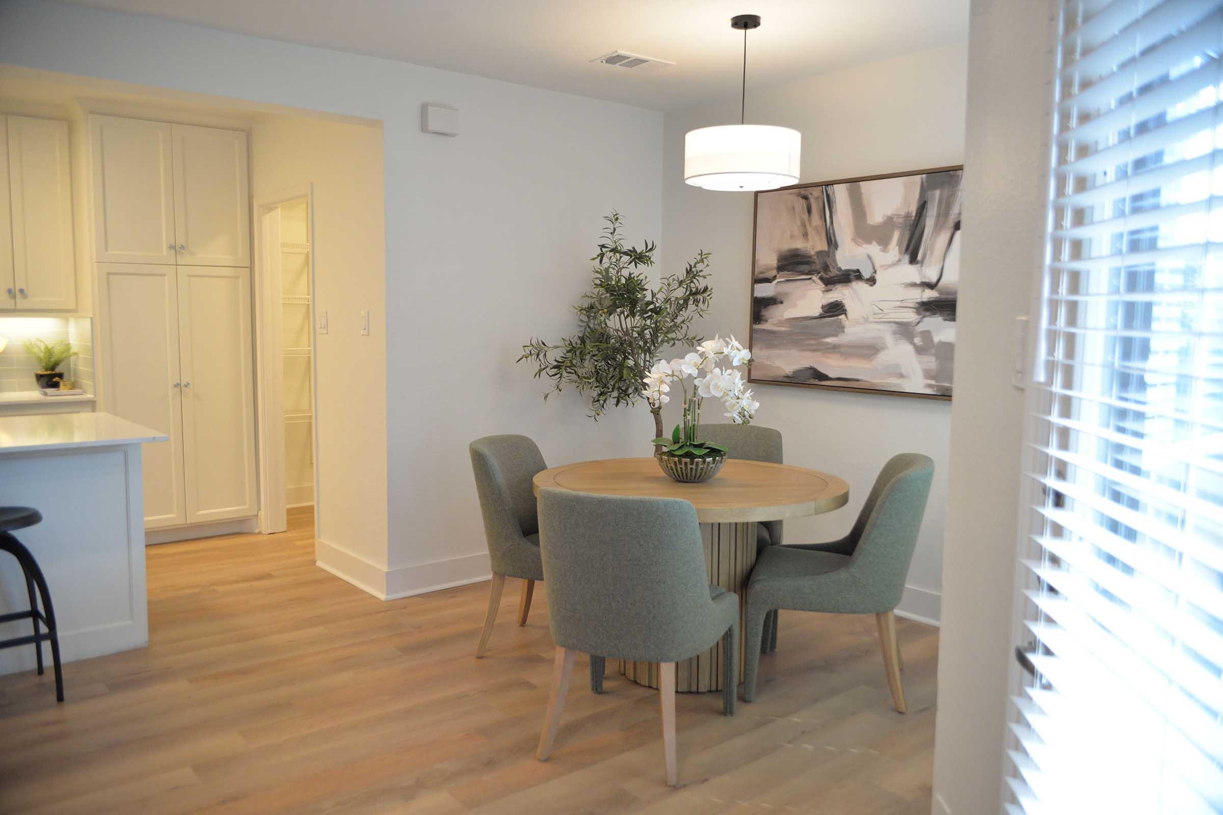 A modern dining area featuring a round wooden table with four upholstered chairs, a vase of white orchids, and a large abstract painting on the wall. Natural light streams in through a window with blinds, and a kitchen area with white cabinetry is visible in the background.