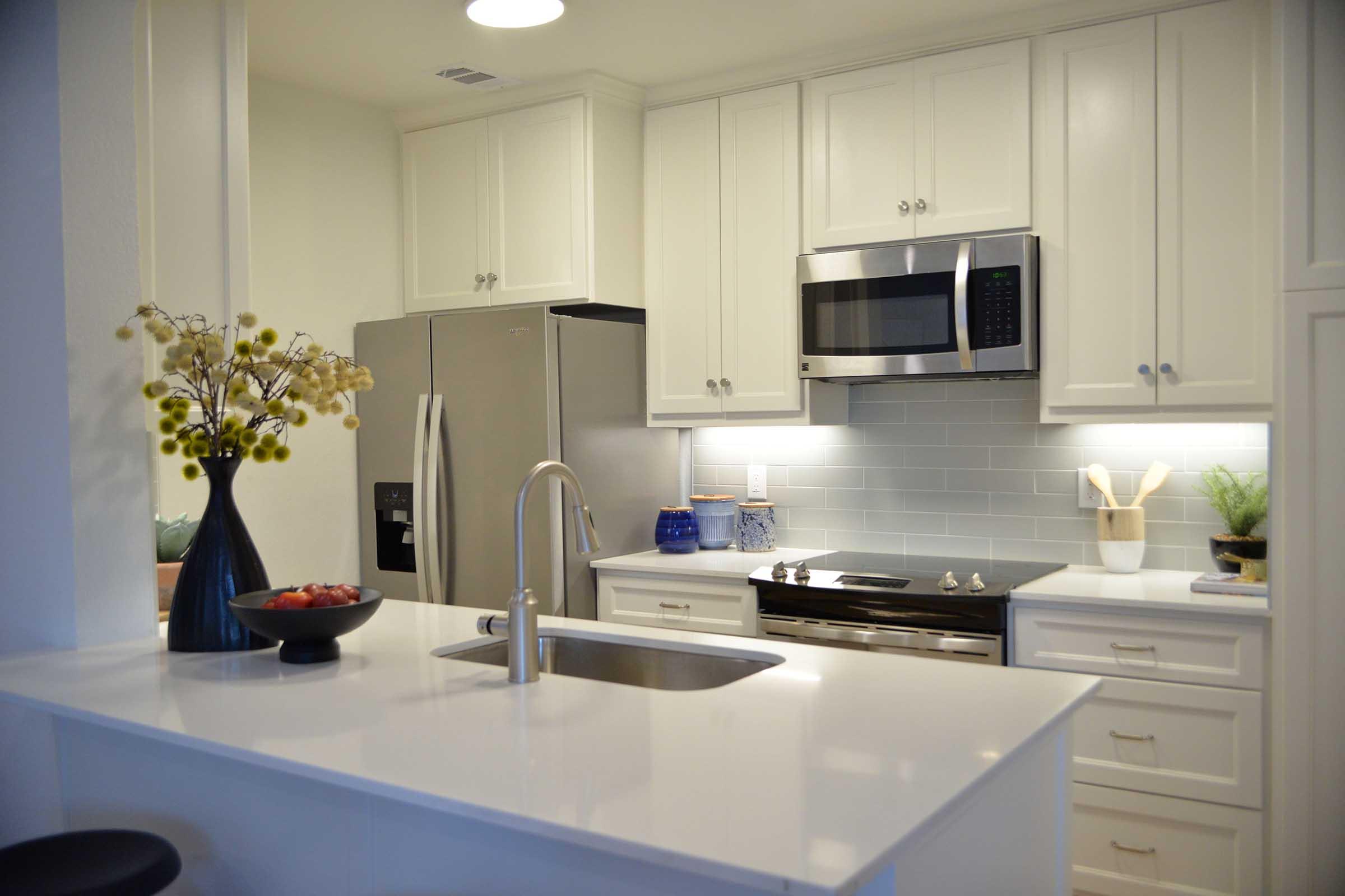 Modern kitchen featuring white cabinetry, stainless steel appliances, and a sleek countertop. A silver refrigerator and microwave are visible, along with a stove. A vase with decorative flowers and a bowl of fruit are placed on the counter, complemented by a subtle backsplash and organized utensils.