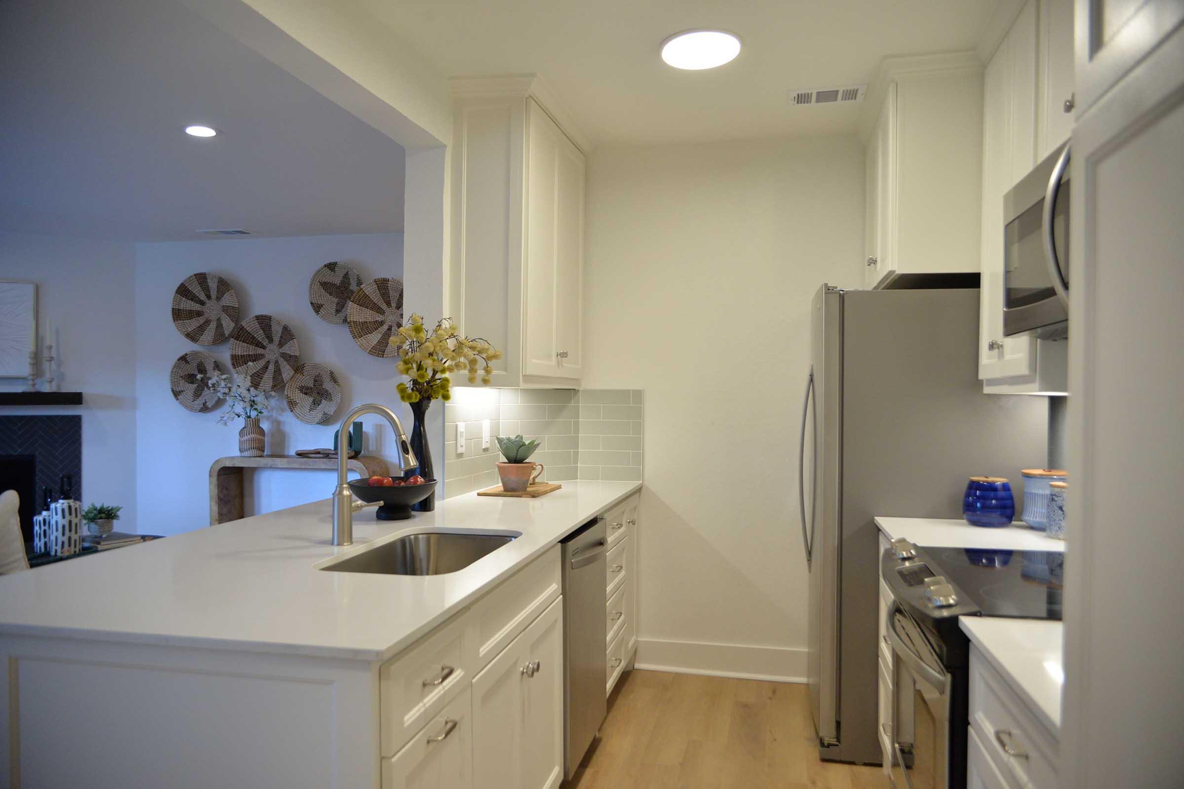 A modern kitchen featuring white cabinetry and a stainless steel refrigerator. The countertop is light-colored with a sink and a small arrangement of plants. Decorative circular wall hangings are visible in the background beside a fireplace. The overall aesthetic is bright and minimalist.