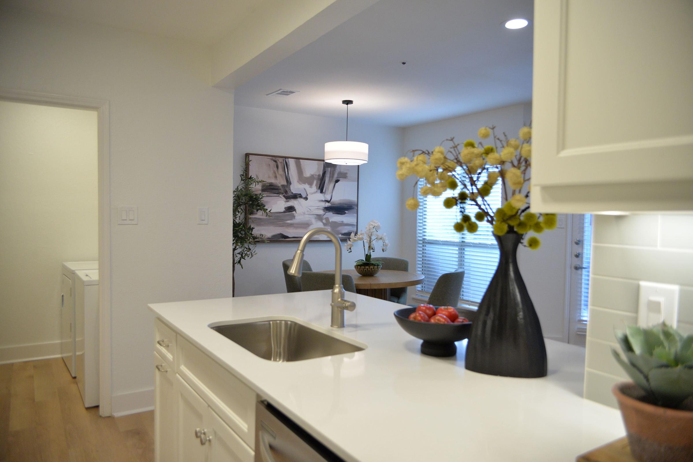 A modern kitchen featuring a sleek white countertop, stainless steel sink, and a black vase with decorative flowers. In the background, a dining area with a round table and a painting on the wall. Light streams in through a window, creating a bright and inviting atmosphere. A laundry area is visible to the left.