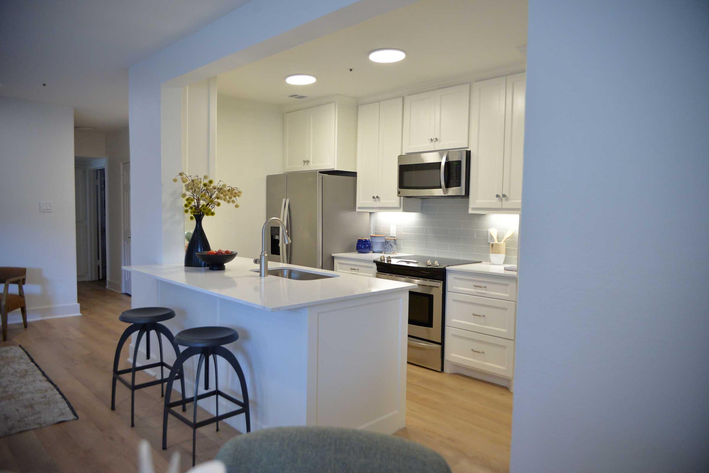 A modern kitchen featuring white cabinetry, stainless steel appliances, and a sleek countertop. Two black stools are positioned at a small island with a vase of flowers. Natural light illuminates the space, creating a bright and inviting atmosphere.
