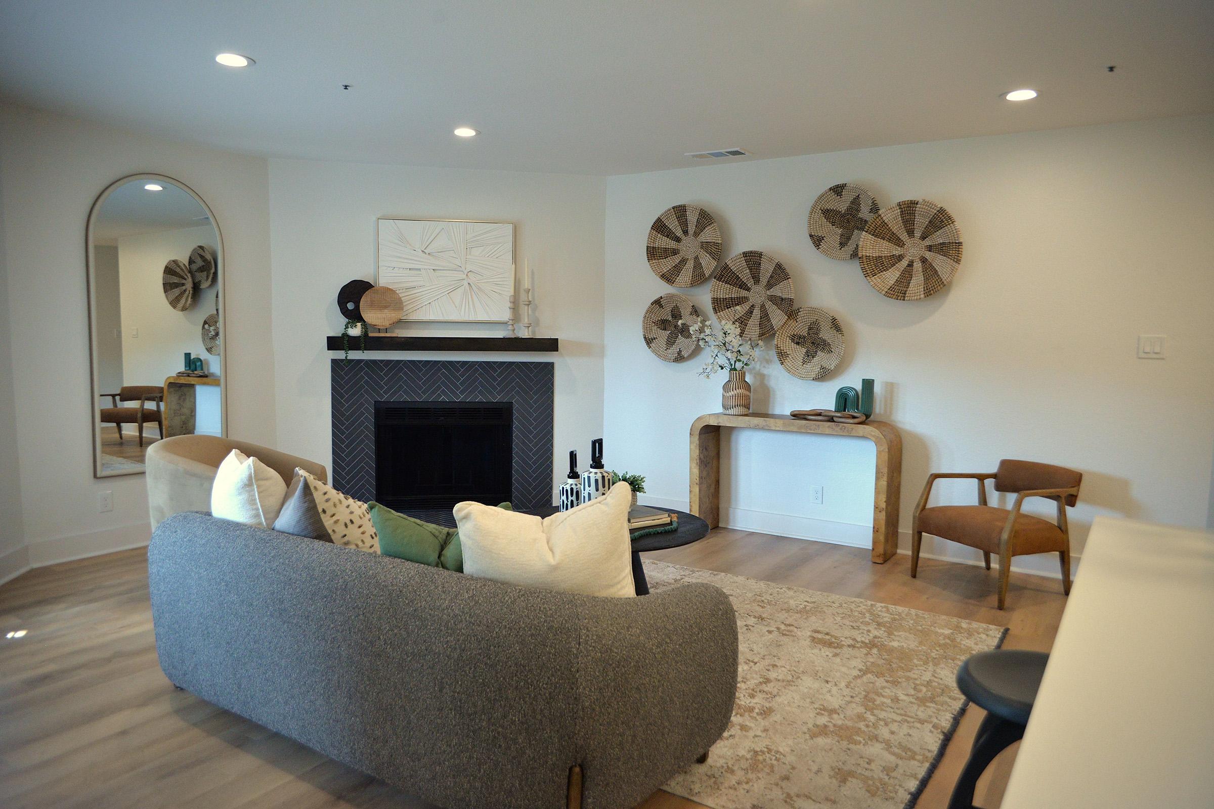 A modern living room featuring a cozy gray sofa with decorative pillows, a fireplace with a dark herringbone pattern, wooden wall art, a light-colored rug, and a stylish console table. The space includes a mirror and a comfortable chair, creating an inviting atmosphere.