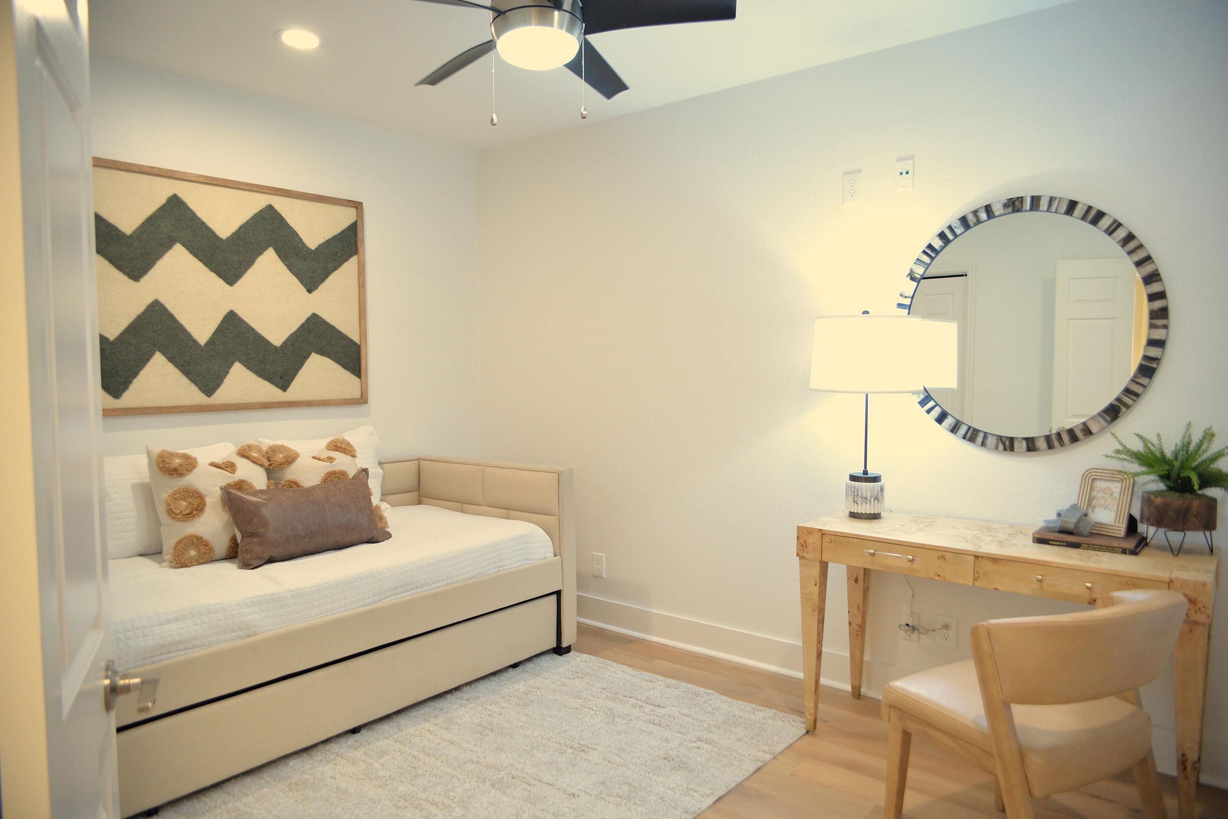 A bright, minimalist bedroom featuring a single daybed with decorative pillows, a round mirror on the wall, a stylish lamp on a wooden desk, and a plant. The decor includes a zigzag-patterned wall art piece, wooden accents, and light-colored walls and flooring, creating a cozy and inviting atmosphere.