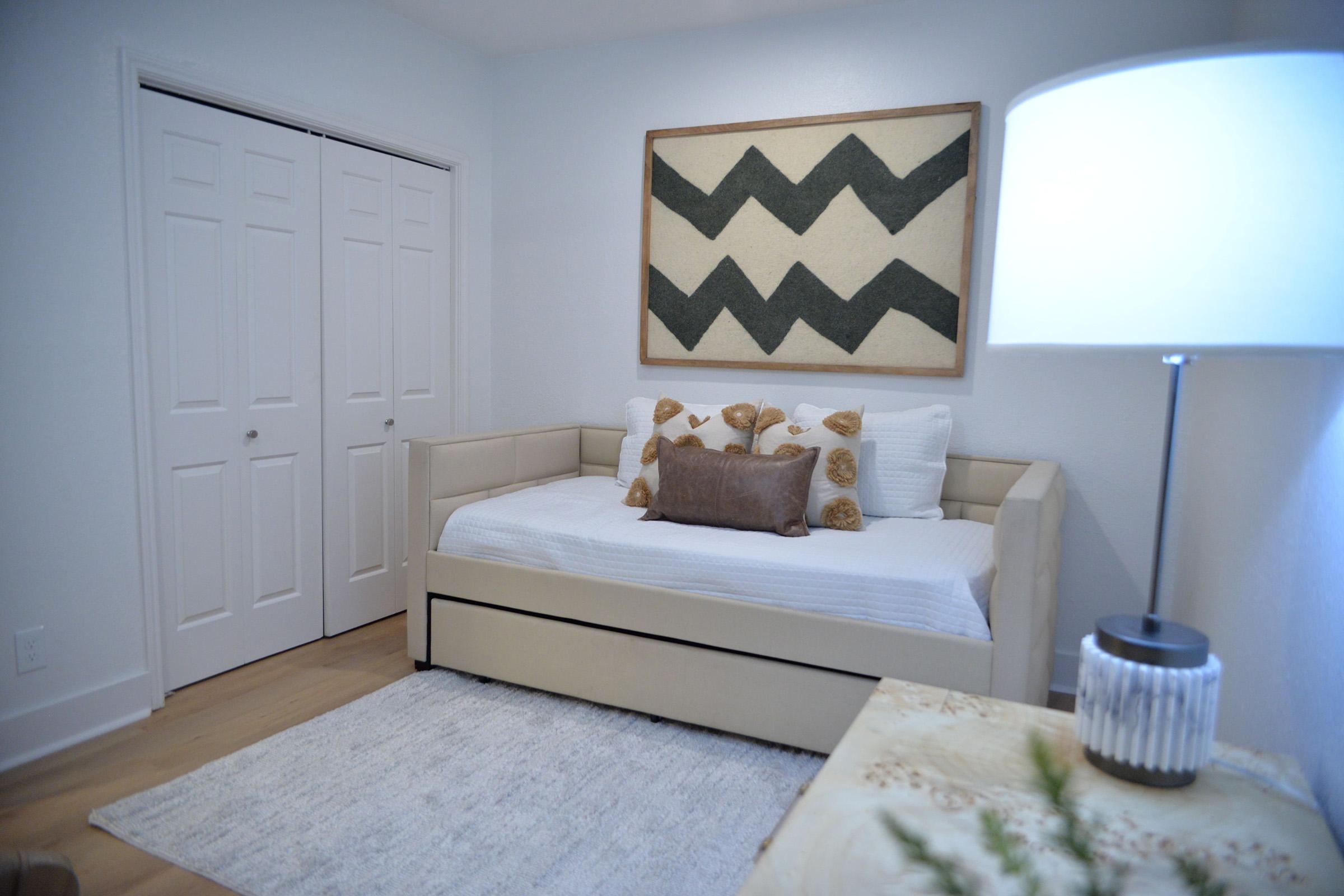 A cozy, modern bedroom featuring a daybed with decorative pillows, a minimalist lamp, and a textured wall art piece with a zigzag pattern. The room has light-colored walls, wooden flooring, and a soft area rug, creating a serene and inviting atmosphere. Two closet doors are visible in the background.