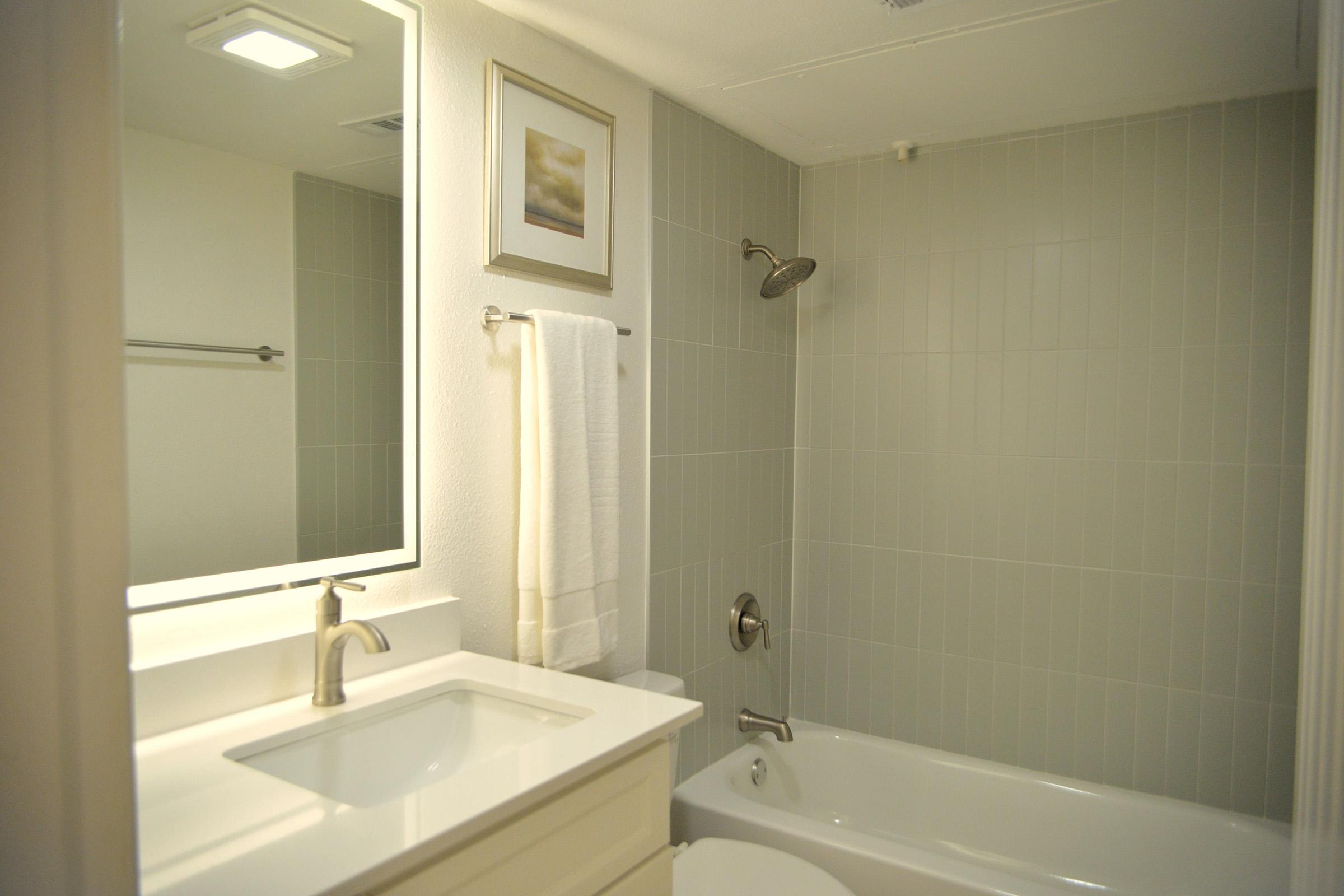 A modern bathroom featuring gray tiled walls, a bathtub with a shower, a white sink with a faucet, a large mirror above the sink, and a towel hanging on a towel rack. A framed picture is mounted on the wall, adding a touch of decor to the space. Soft lighting enhances the clean, inviting atmosphere.