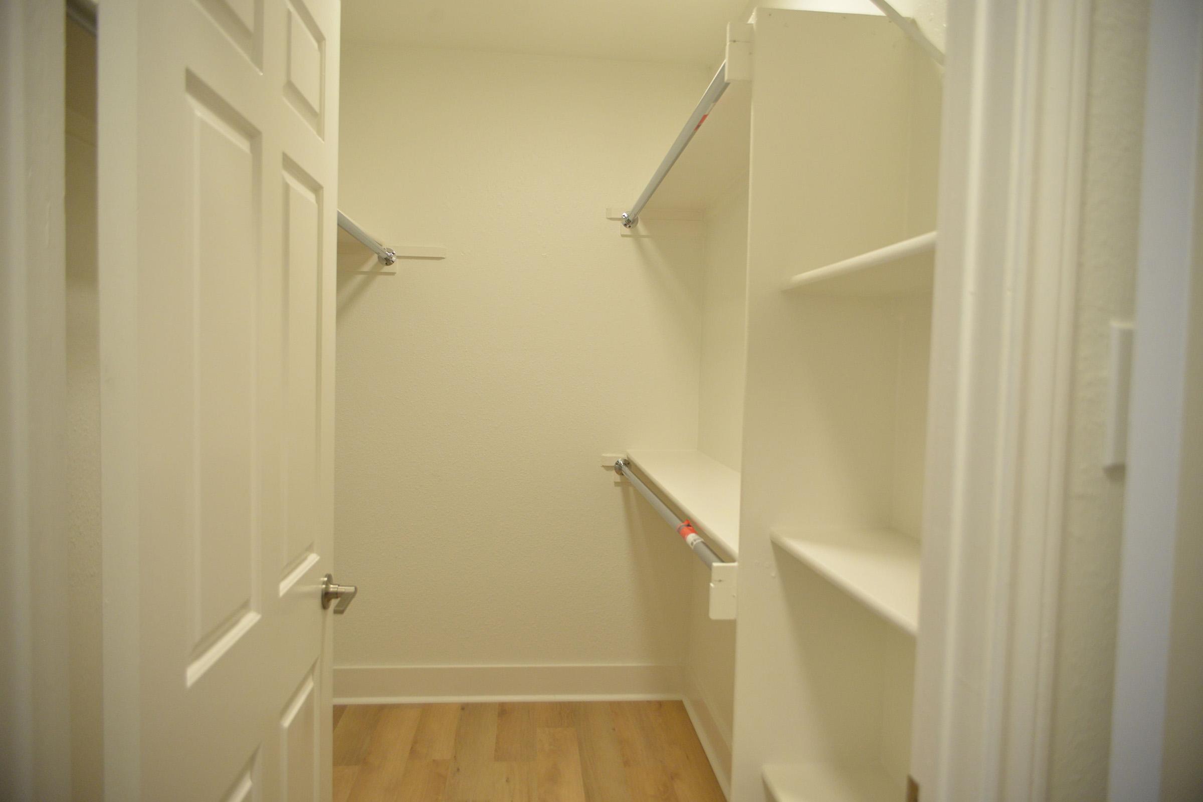 A bright, empty closet with white walls and wooden flooring. There are two horizontal rods for hanging clothes and several shelves for storage, creating a simple and organized space. The door is closed, and the overall atmosphere is clean and spacious.