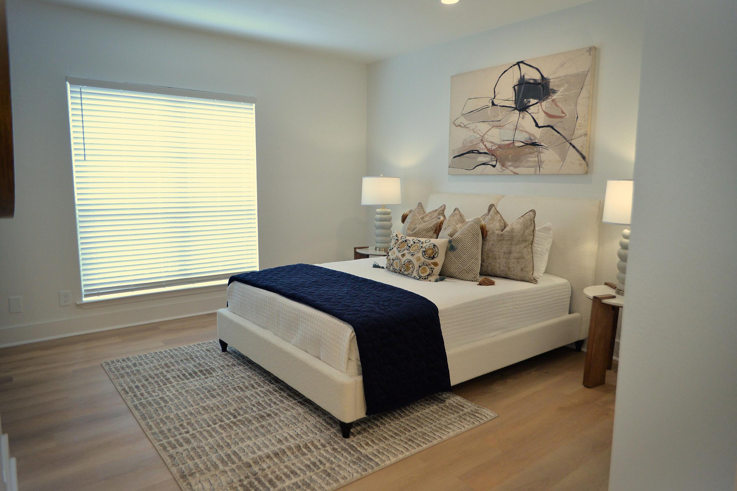 A cozy, modern bedroom featuring a neatly made bed with decorative pillows and a dark blue throw blanket. Soft lighting from two bedside lamps illuminates the room, while a piece of abstract art hangs above the bed. A large window with blinds allows natural light to filter in, enhancing the serene atmosphere.