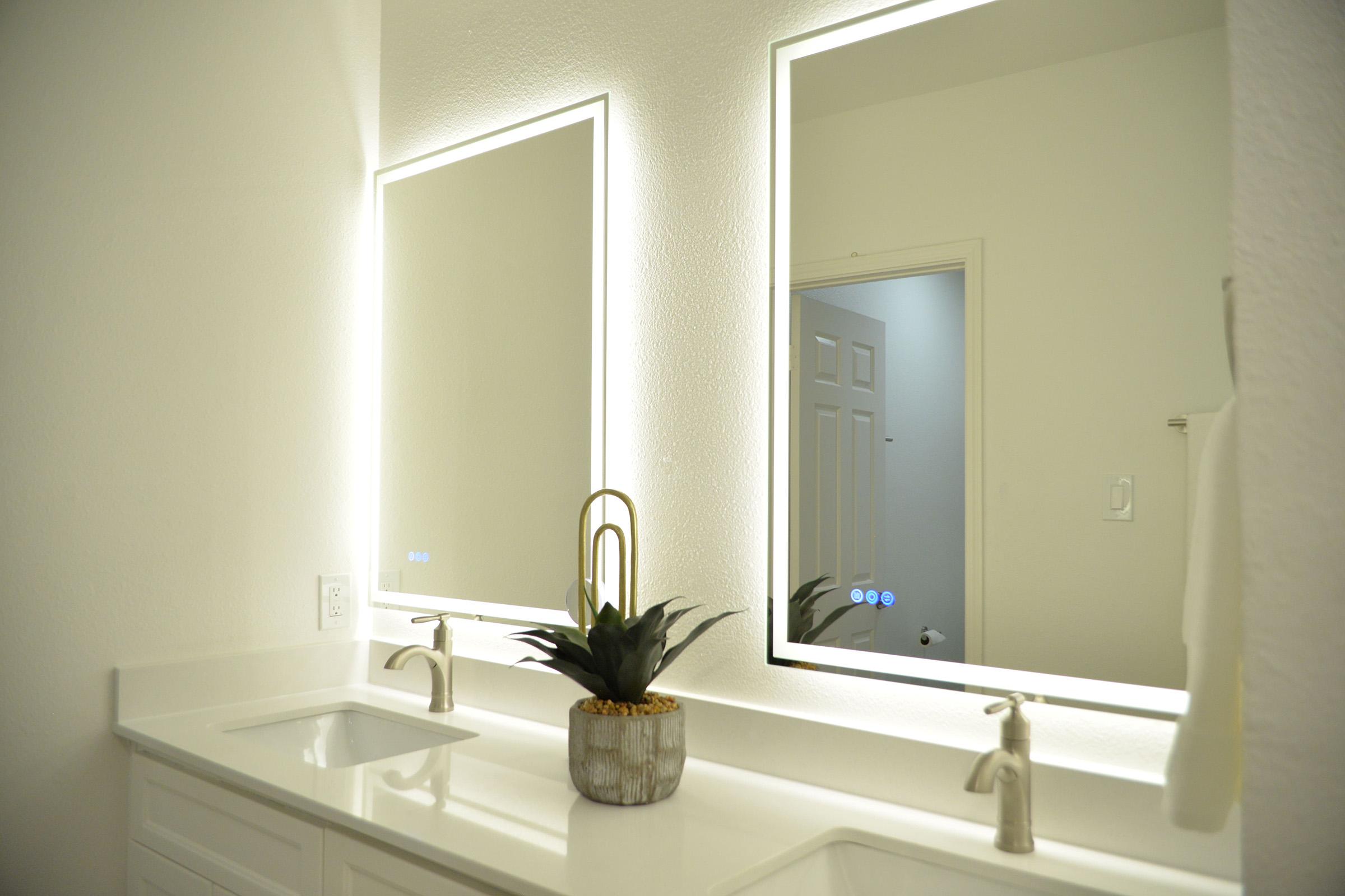 A modern bathroom featuring two illuminated mirrors above a white double sink vanity. A small potted plant sits between the sinks. The walls are painted a light color, and a door to another room is visible in the background. The overall design is clean and contemporary.