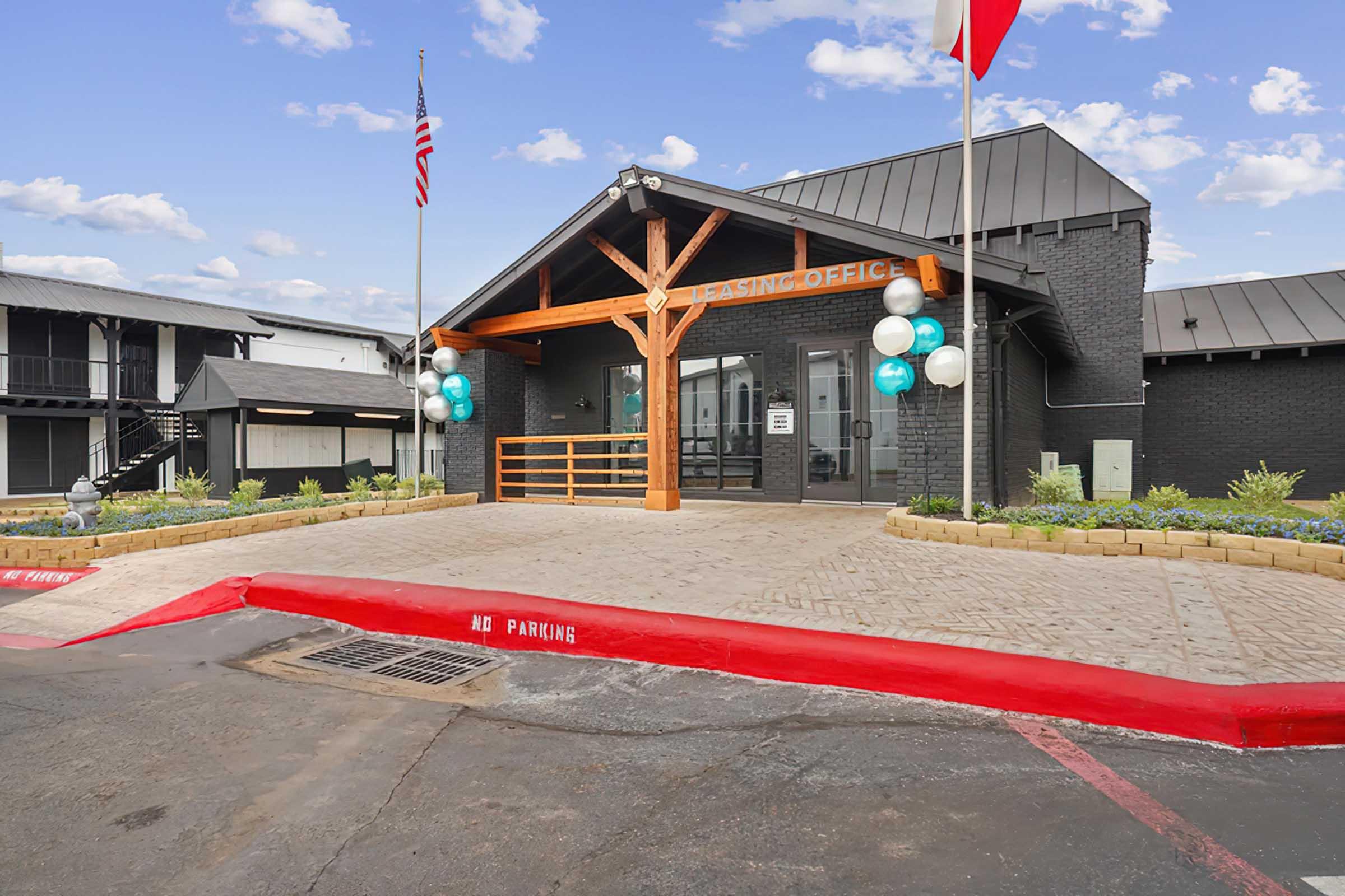 Exterior view of a leasing office with a wooden entryway, flags, and decorative balloons. The building features a modern design with a black exterior, surrounded by well-maintained landscaping. A red-painted curb marked "No Parking" is visible in front of the office, with a clear blue sky in the background.