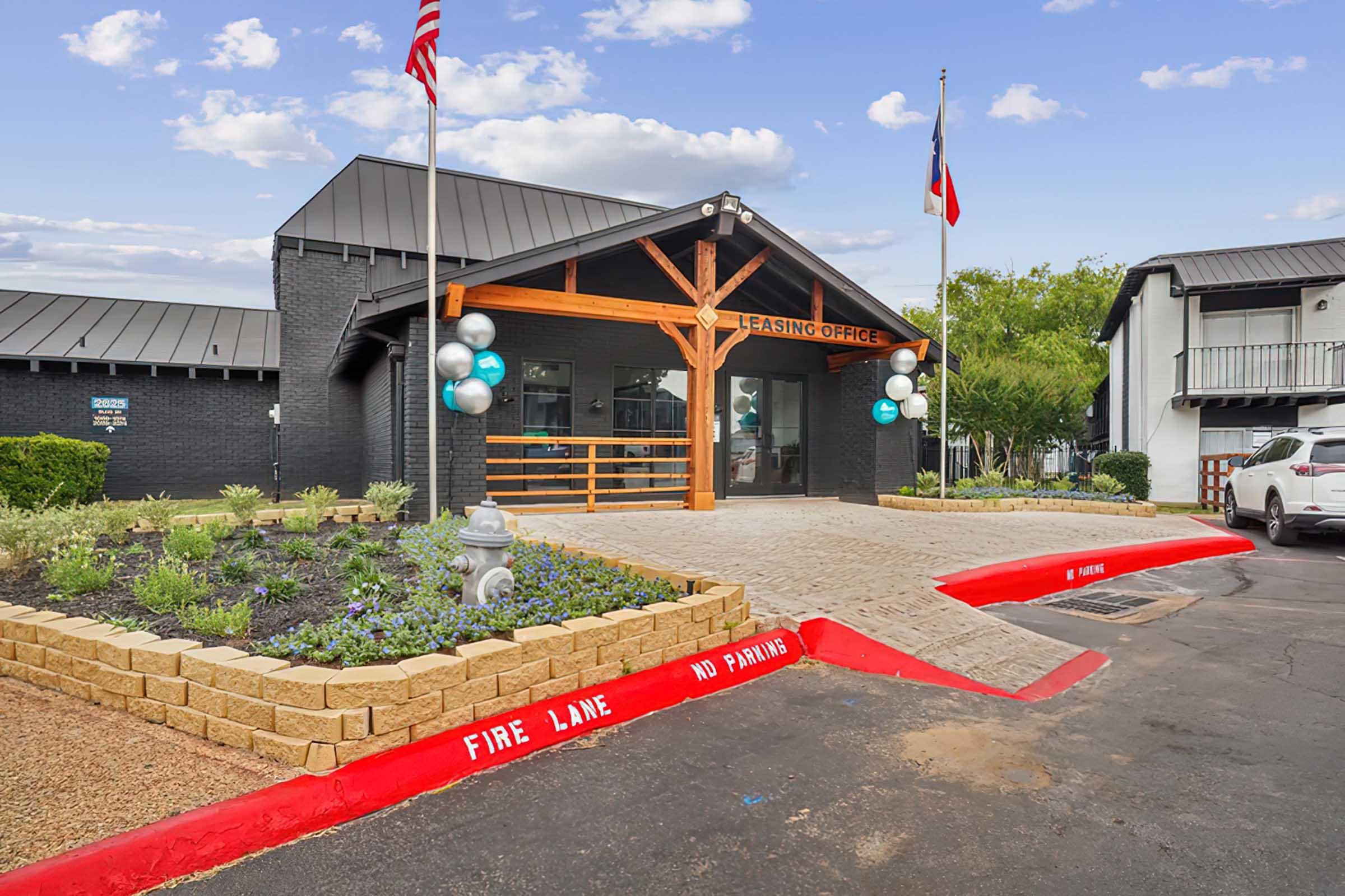 A leasing office entrance featuring a modern design with a wooden beam structure, adorned with blue and white balloons. The pathway is paved with stones, surrounded by shrubs and flowers. There are two flags displayed, and a sign indicates a fire lane with no parking in the area.