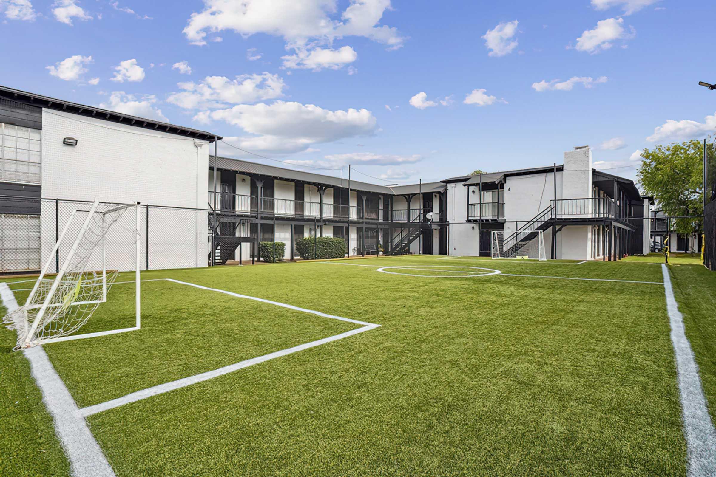 A well-maintained artificial turf soccer field with goalposts, surrounded by a multi-story building with balconies. The sky is partly cloudy, creating a pleasant atmosphere for outdoor activities.