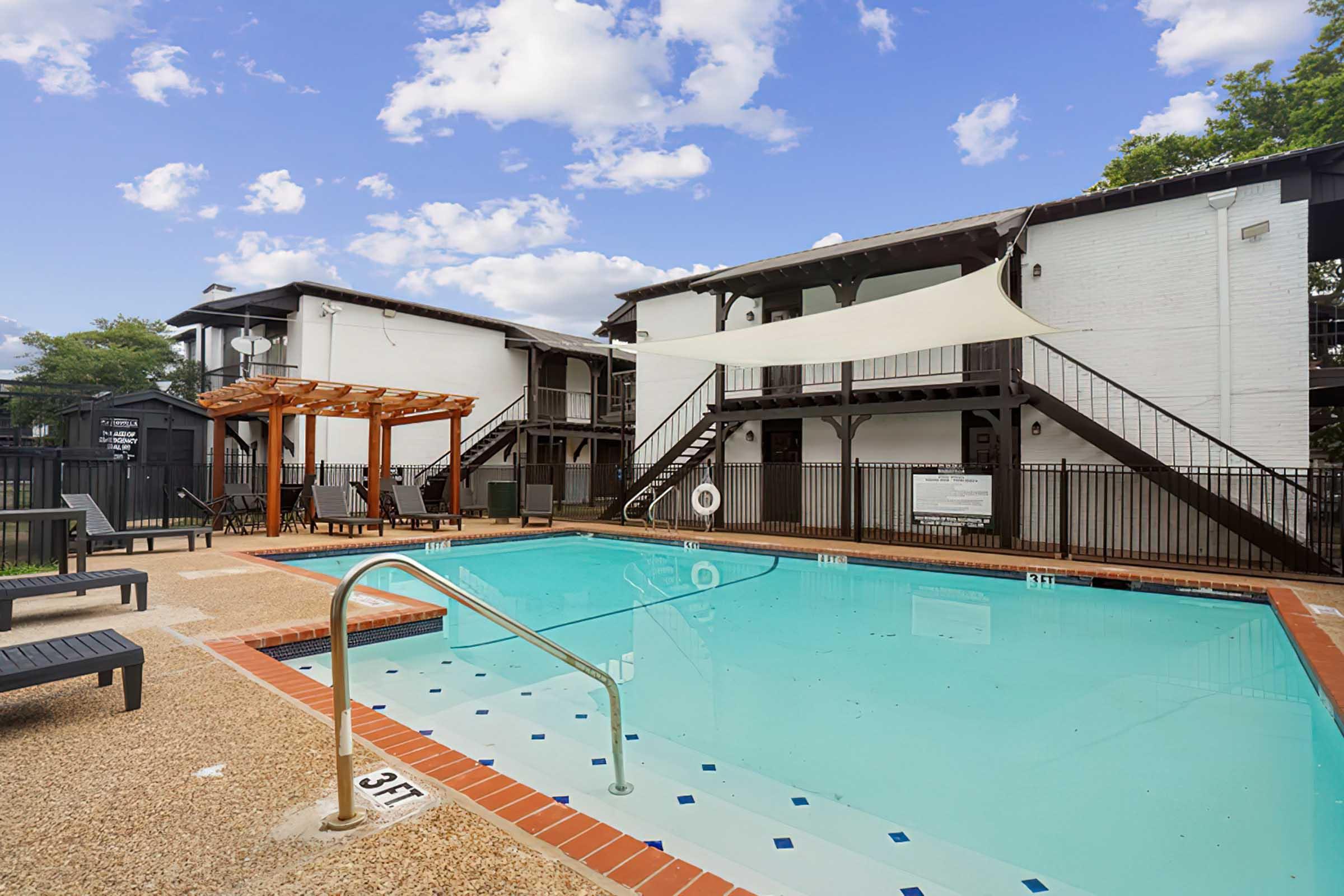 A refreshing swimming pool surrounded by lounge chairs and a wooden pergola. The area features two stylish buildings with balconies, under a bright blue sky with fluffy clouds. Safety signage and a life preserver are visible near the pool area.