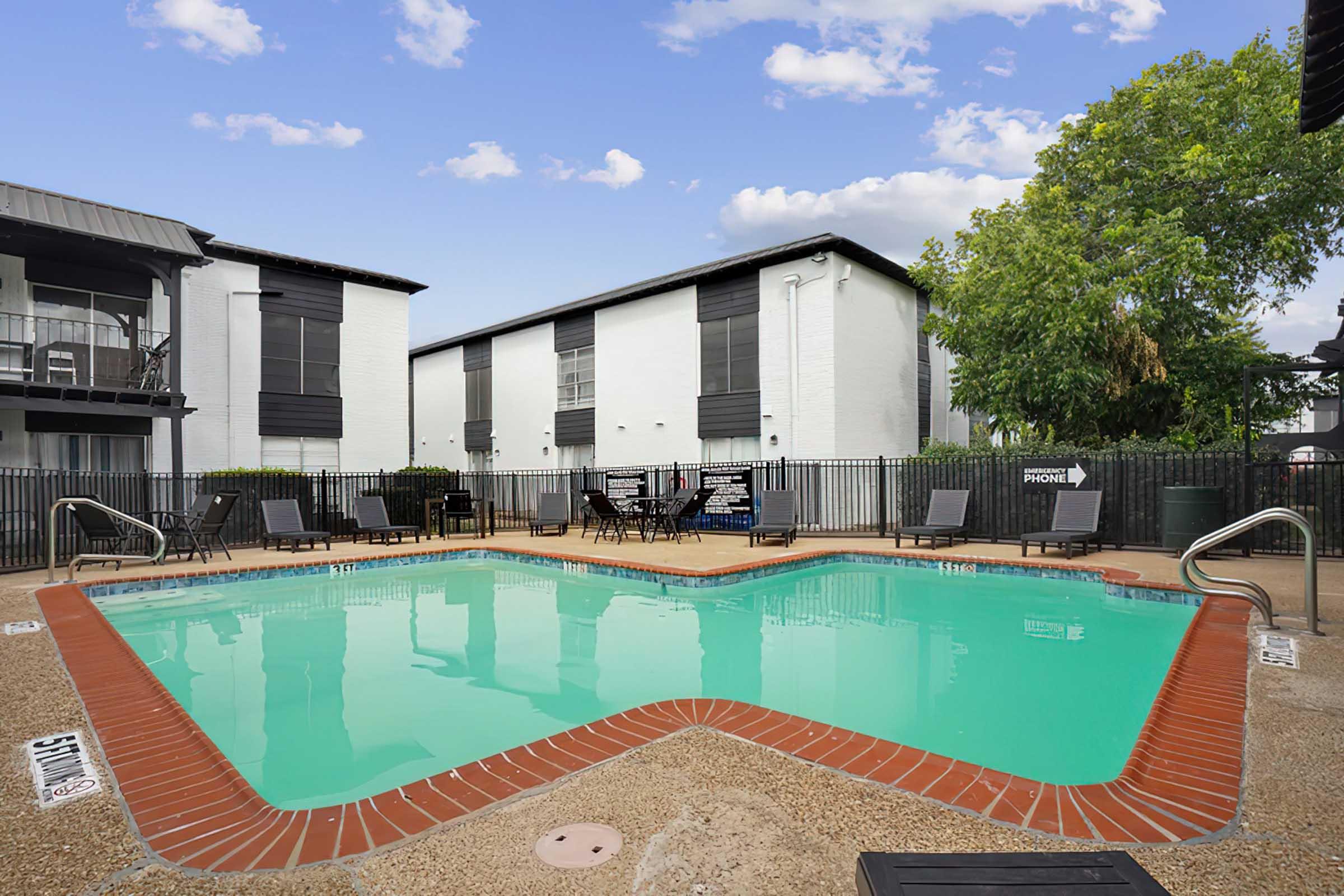 A swimming pool with a turquoise surface and a textured concrete surround. Green trees are visible nearby, along with lounge chairs and tables placed around the pool. The background features two buildings with modern architecture, and a clear blue sky with scattered clouds.