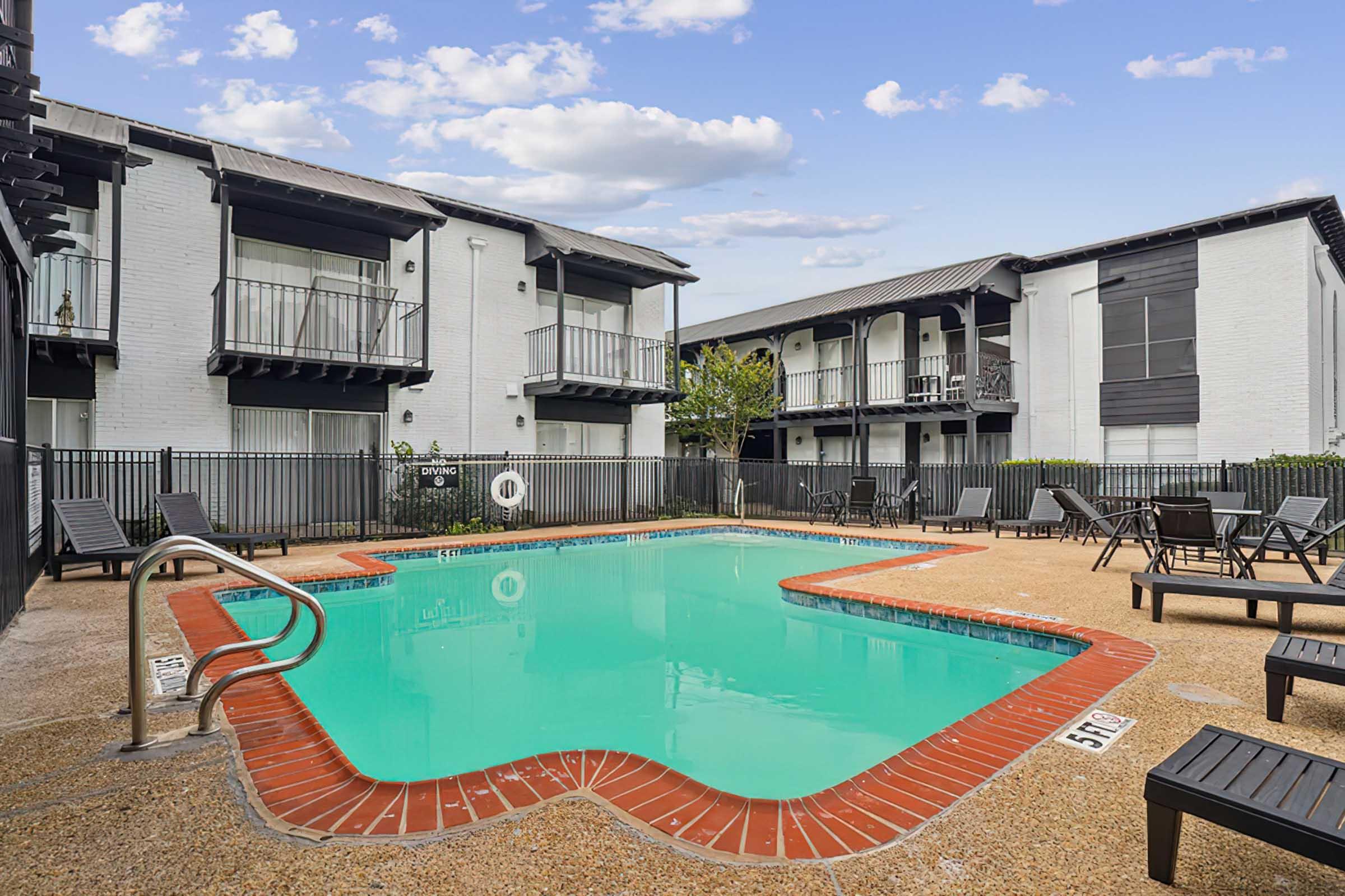 A clear swimming pool surrounded by lounge chairs, situated in a courtyard of a multi-story apartment complex. The pool has a curved shape with a red-brick border. Lush greenery is visible in the background, and several balconies overlook the pool area under a bright blue sky with scattered clouds.