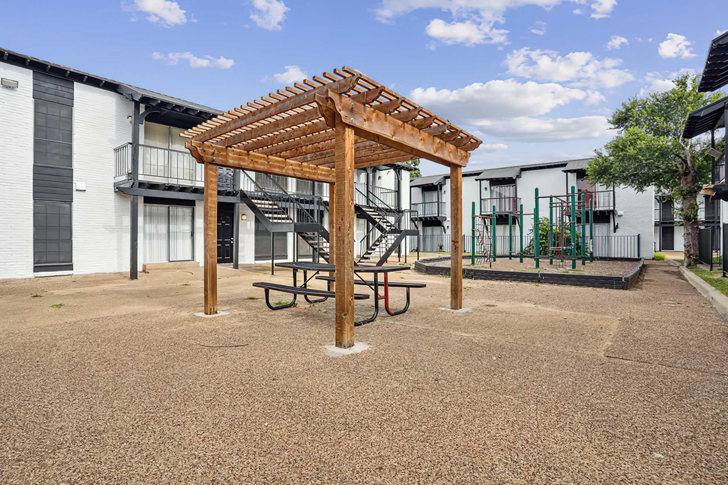 An outdoor area featuring a wooden pergola with a picnic table, surrounded by gravel. In the background, there are two multi-story residential buildings and a playground structure with swings and climbing equipment. The sky is partly cloudy, adding a bright ambiance to the scene.