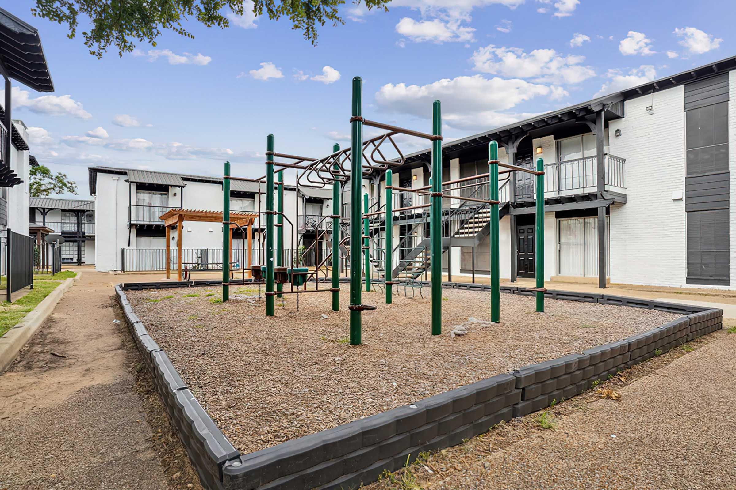 Playground area with climbing equipment surrounded by gravel, located in an apartment complex. Green metal structures for children’s activities are visible, with residential buildings in the background. Clear sky and greenery are present, creating a welcoming outdoor space.