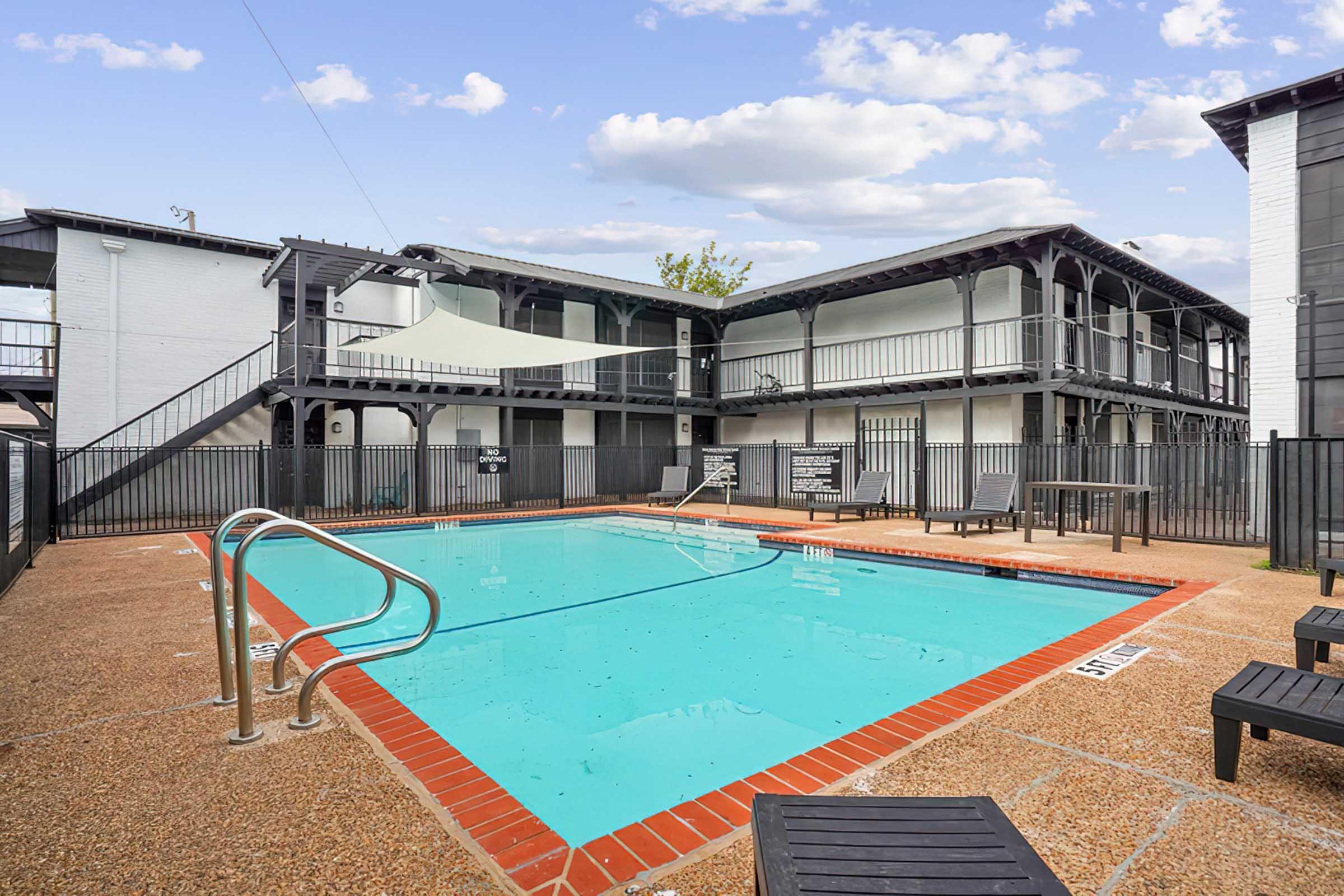 A clear blue swimming pool surrounded by a patio area, with lounge chairs and a protective fence. In the background, a two-story building features balconies. The setting is bright and inviting, under a partly cloudy sky.