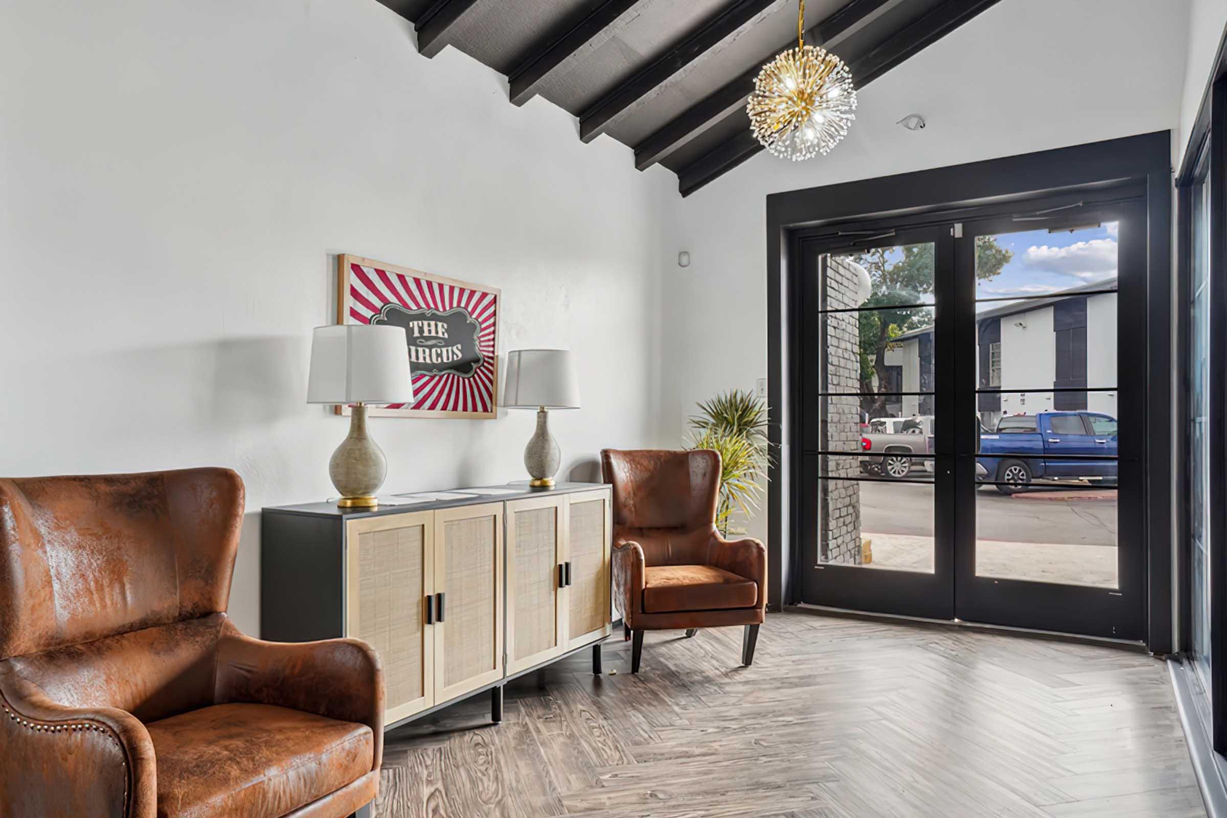 A spacious entryway featuring two brown leather armchairs, a stylish side table with two lamps, and a decorative artwork on the wall. Large glass doors provide a view of the outside, showcasing a parking area. The ceiling has exposed beams, and the floor is finished with a wood-like pattern.
