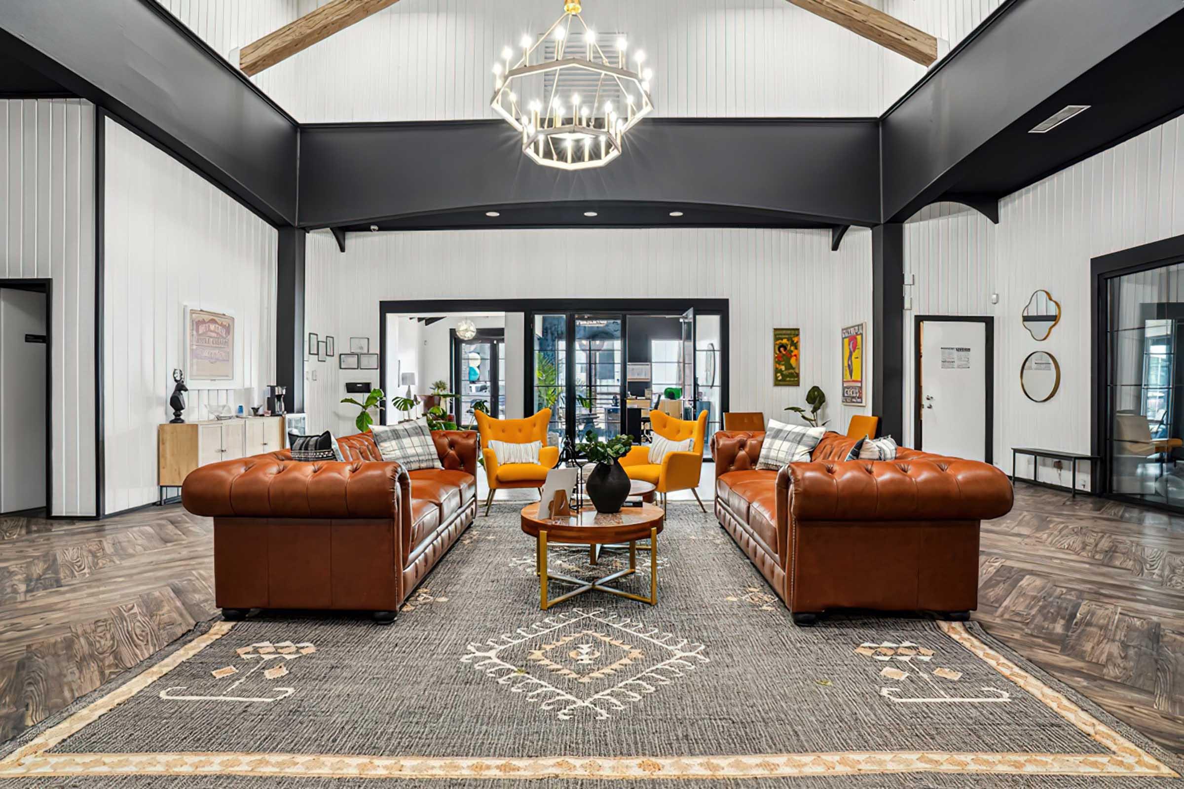 A stylish interior of a modern lounge featuring two brown leather sofas facing each other, a circular coffee table in the center, and vibrant yellow armchairs. The space is adorned with wooden beams, decorative artwork, and greenery, creating a warm and inviting atmosphere. A large chandelier hangs overhead.