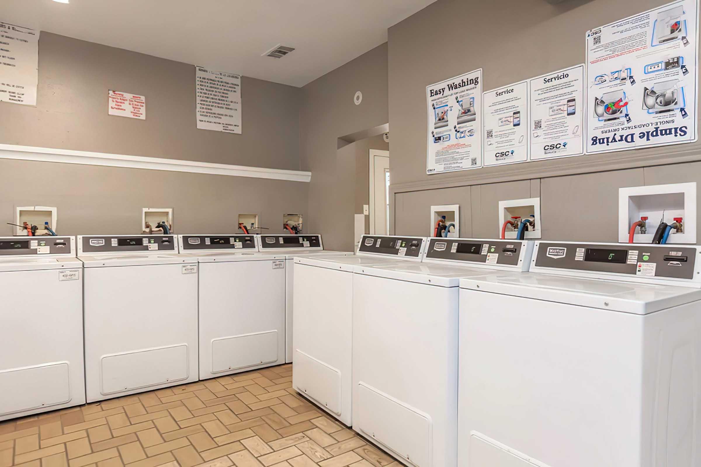 A bright and clean laundromat featuring several white washing machines lined up against a light gray wall. Instruction signs and service posters are visible on the walls. The floor is tiled with light-colored tiles, creating a neat and organized environment for laundry services.