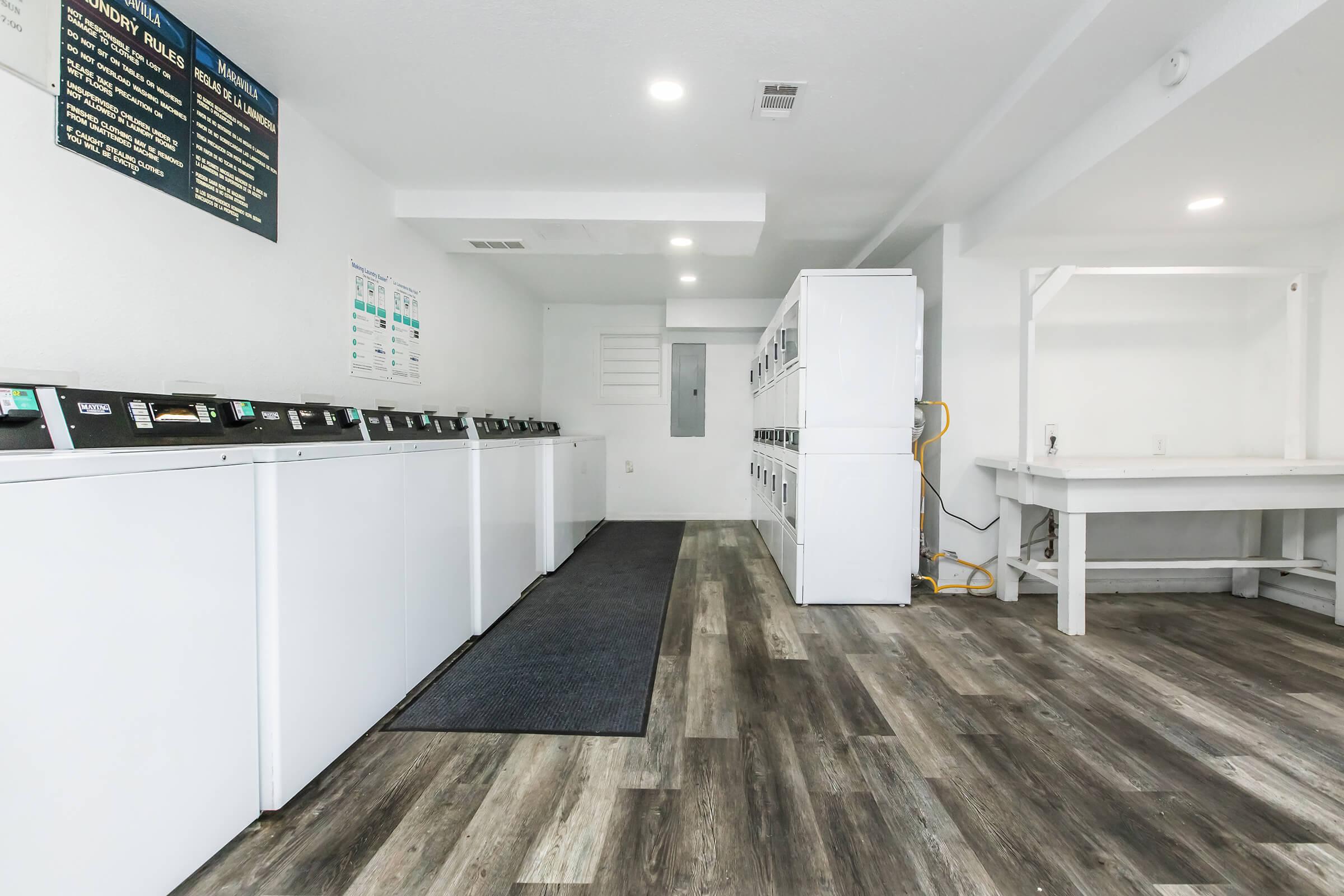 A clean, well-lit laundry room featuring several white washing machines lined up against the wall, a gray mat on the floor, and a row of lockers or dryers. The walls are painted white, and there are informational posters displayed. A folding table is seen in the corner of the space.