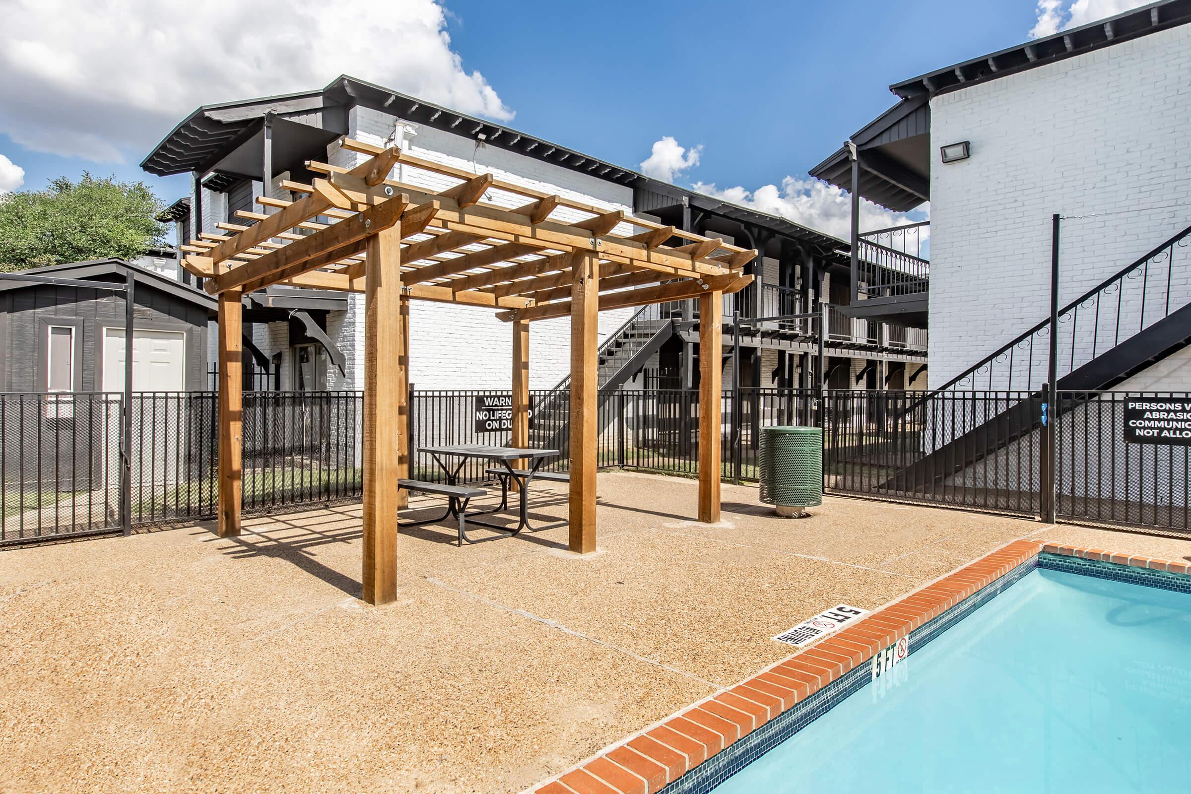A wooden pergola alongside a swimming pool, with a small table and chairs underneath. The pool is surrounded by a fence, and there are white apartment buildings in the background against a partly cloudy sky.