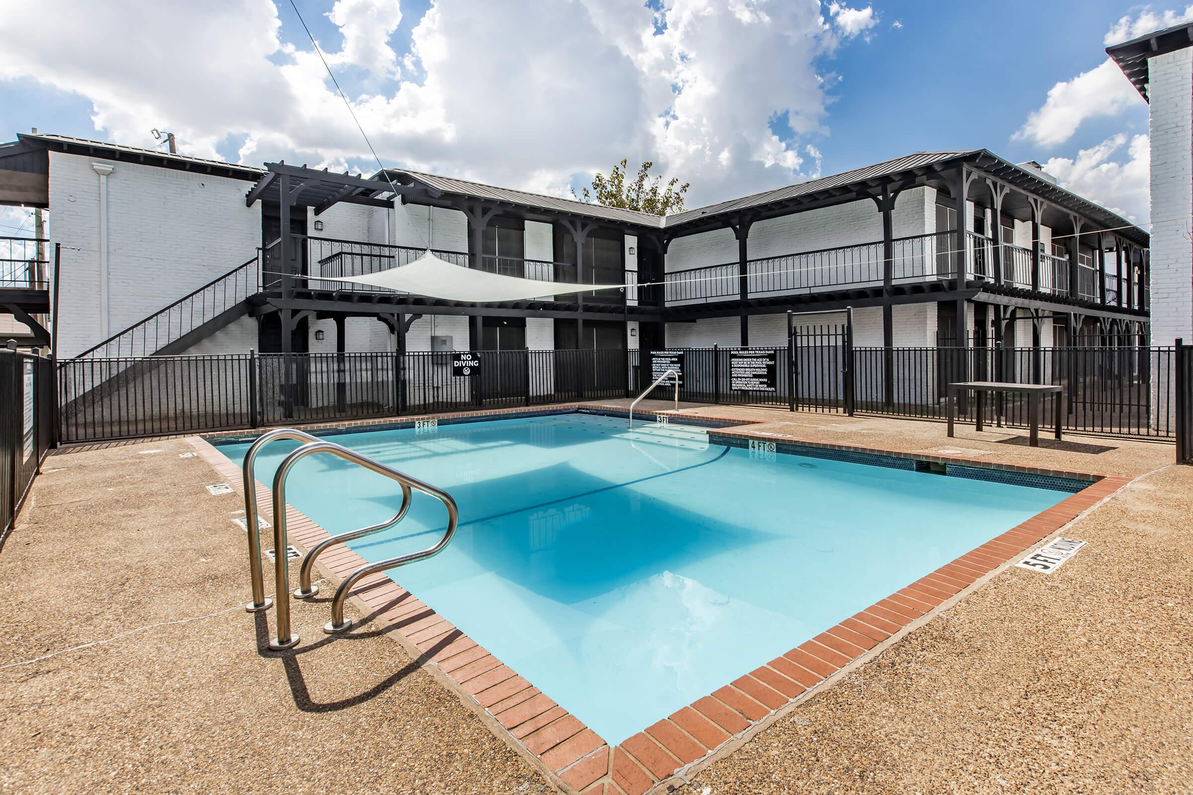 A well-maintained outdoor swimming pool surrounded by a decorative fence, situated near multi-story apartments. The pool has ladder access and is framed by bright, sunny weather with fluffy clouds in the sky. A shaded area is visible above the pool, enhancing the inviting atmosphere.